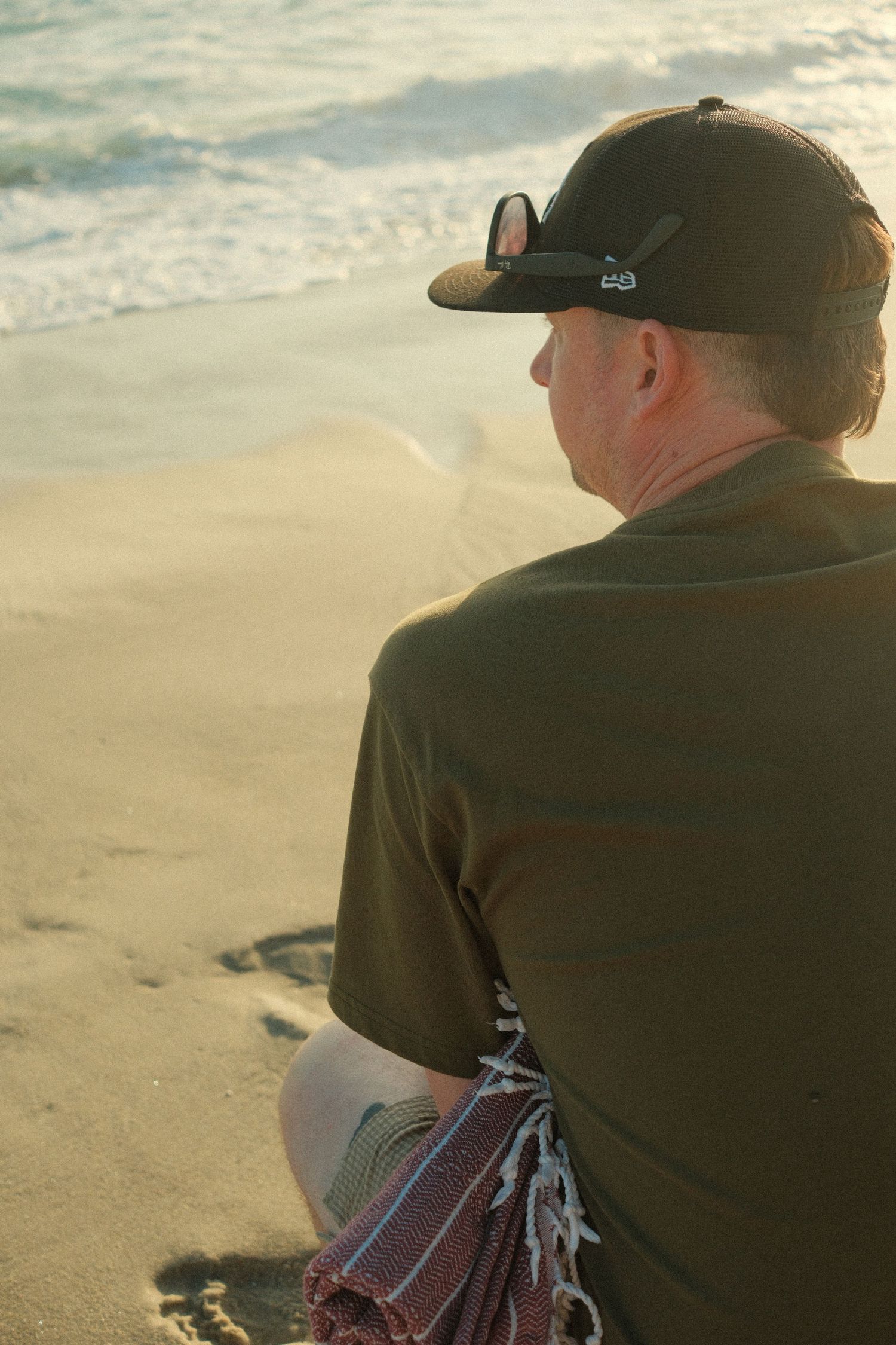 Man in dark green shirt and black cap sits on sandy beach gazing at ocean waves during golden hour.