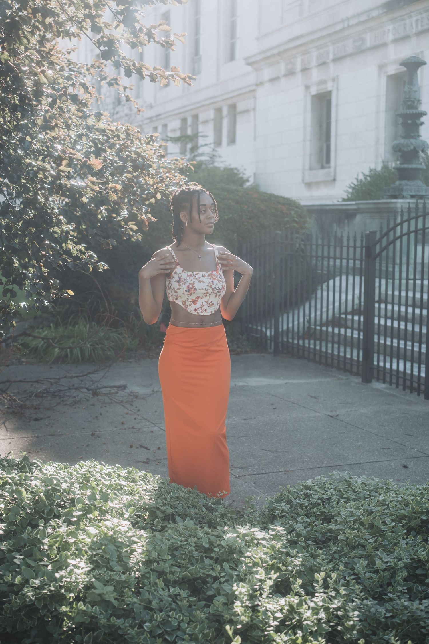 Woman in floral crop top and orange maxi skirt posing outdoors near lush greenery and a white building.