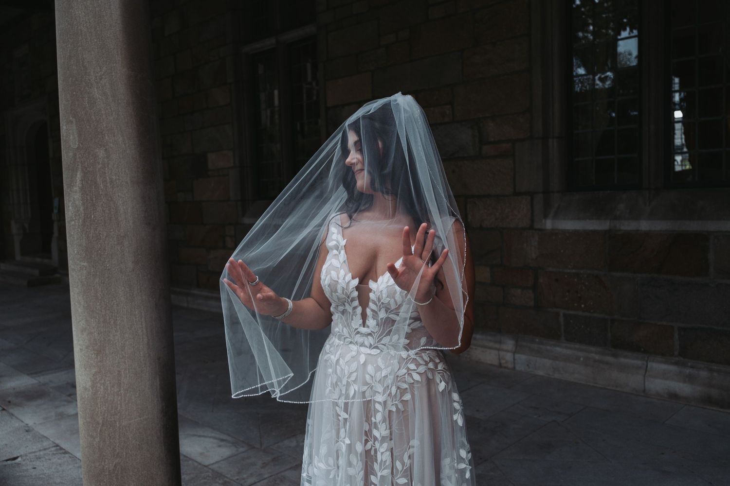 A person in an elegant lace wedding dress poses in a dimly lit architectural setting.