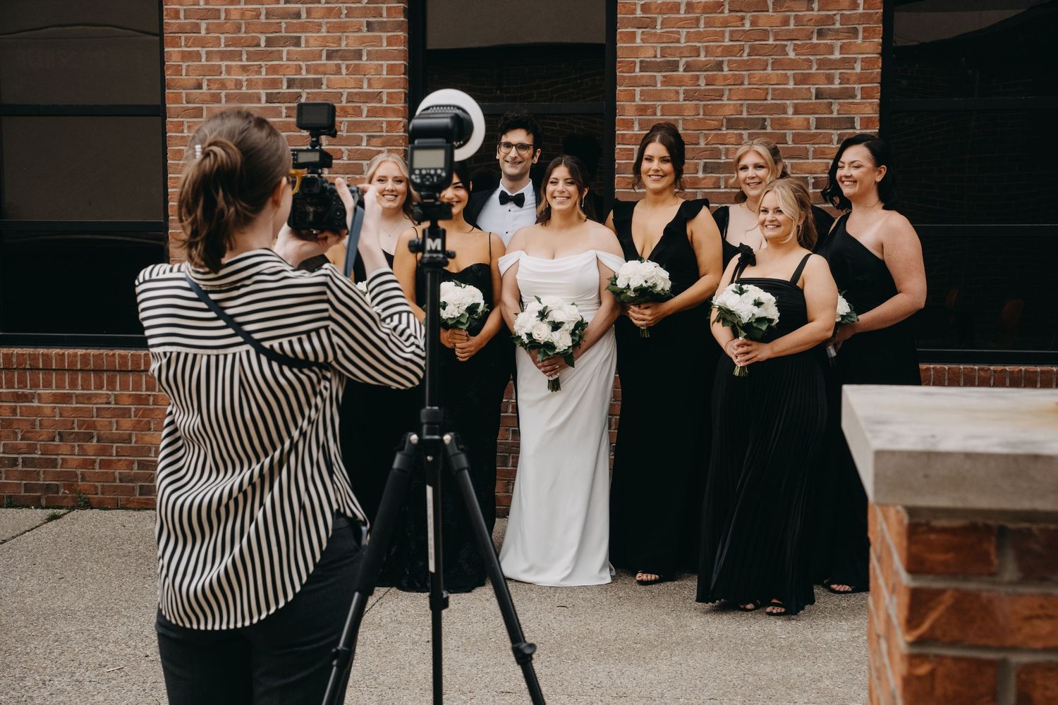 Photographer capturing a wedding party with bridesmaids in black dresses and bride holding white bouquet.