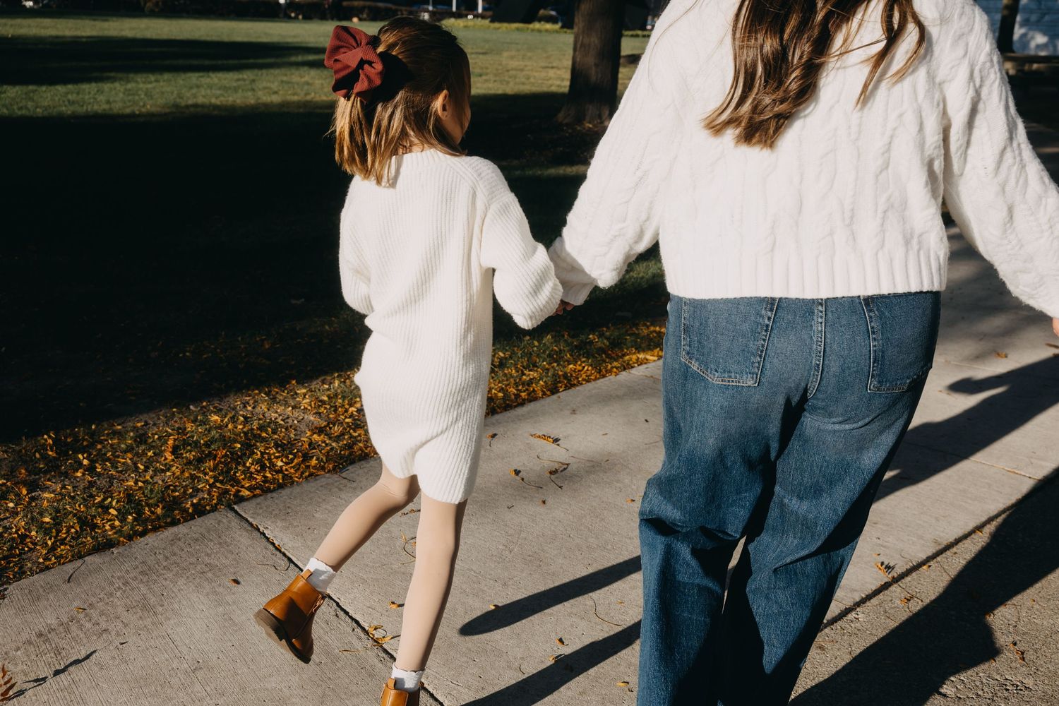 Two girls holding hands walking on a sidewalk outdoors, wearing white sweaters and jeans on a sunny day.