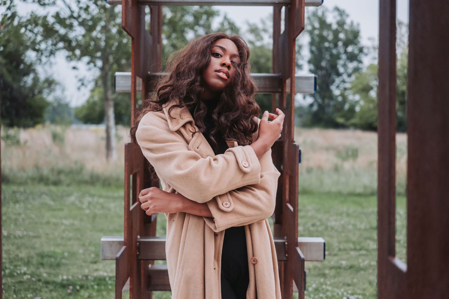 Woman in beige trench coat posing outdoors near wooden structure with lush green field background.