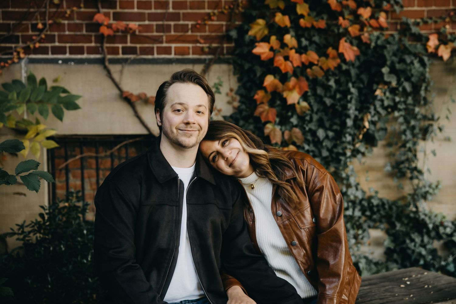 Couple posing together outdoors in autumn, woman resting head on man's shoulder, surrounded by colorful fall foliage.