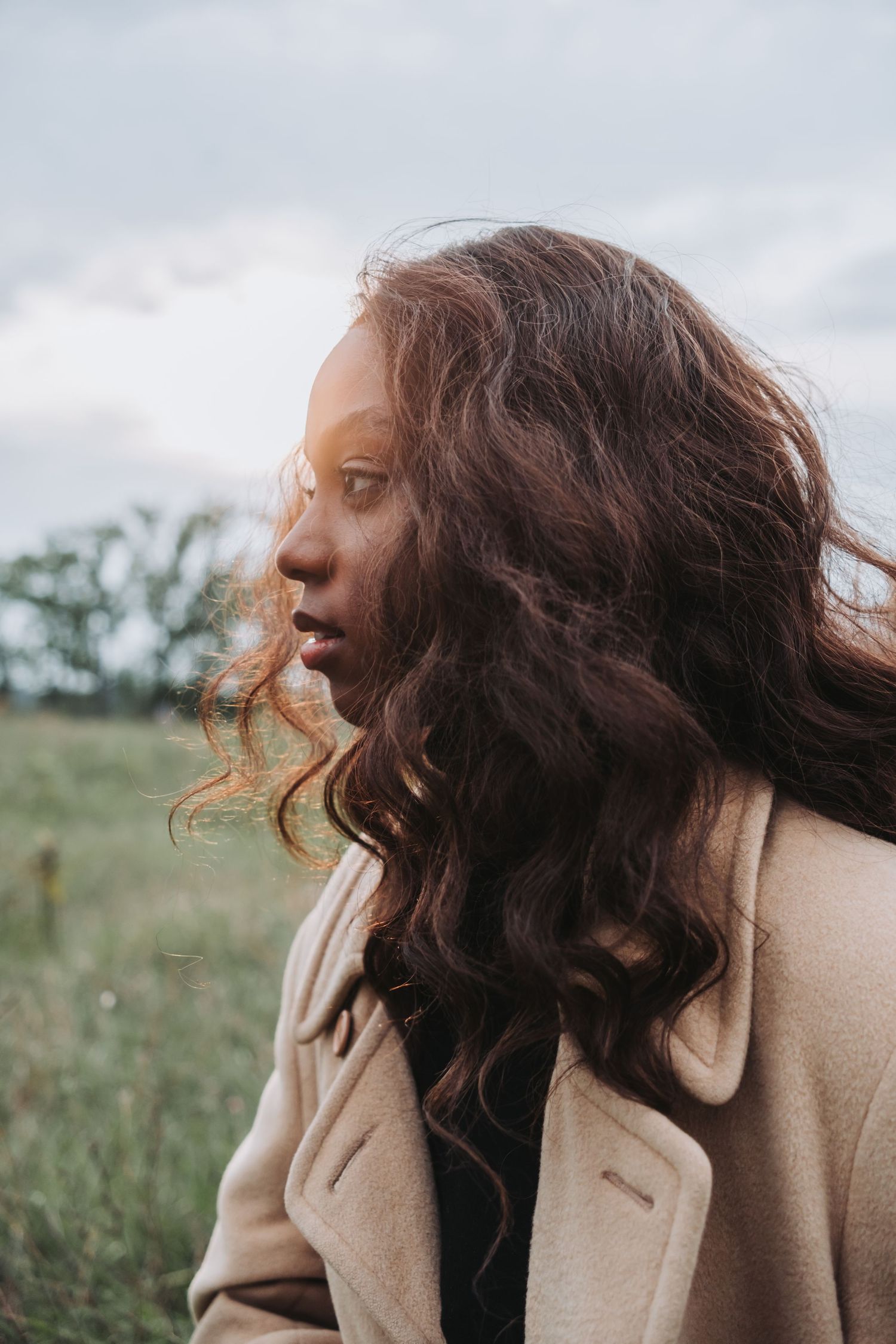 A young Black woman with long curly brown hair stands in a field, wearing a beige coat, her hair blowing in the wind.