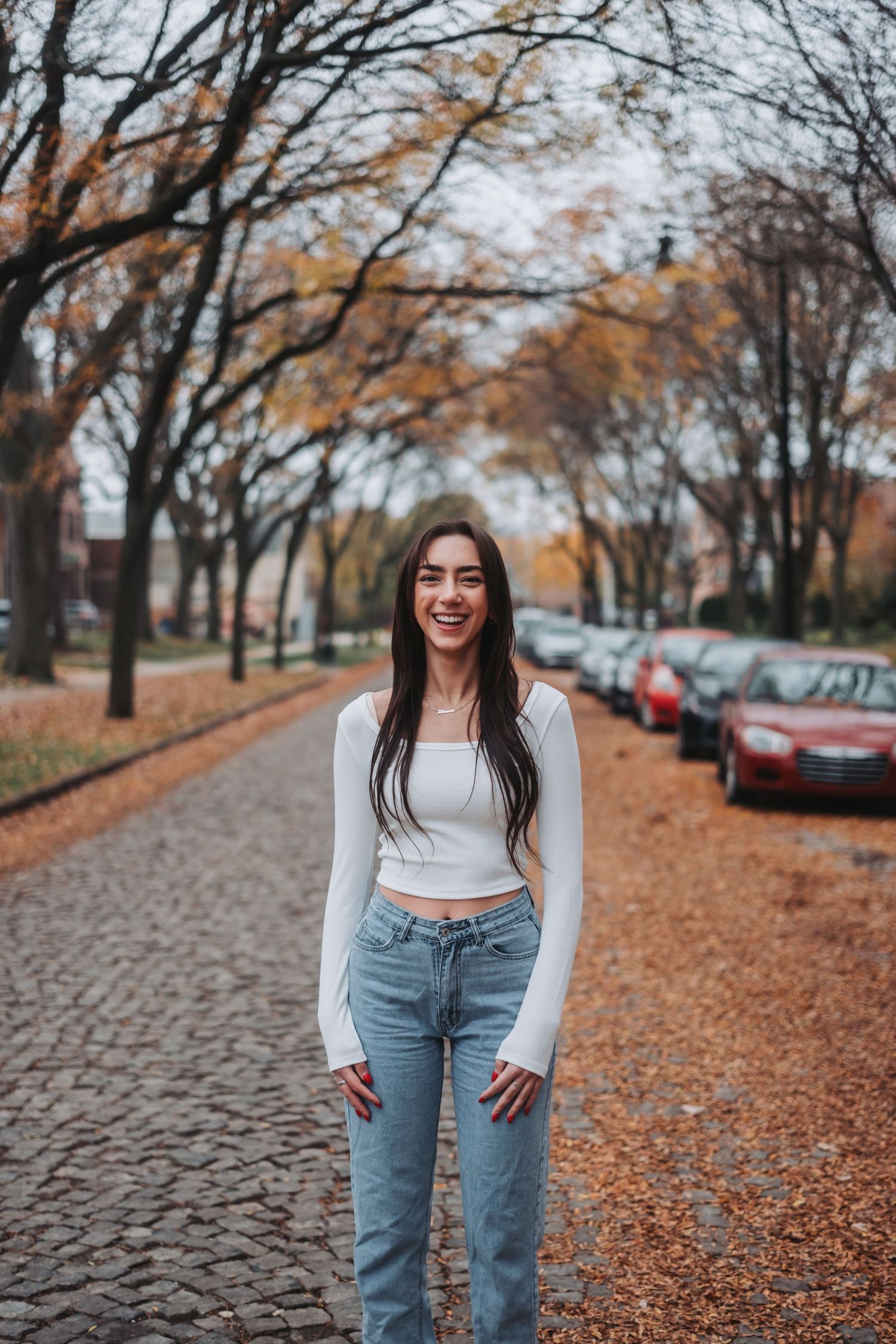 Young woman in white crop top and jeans smiling on a cobblestone path lined with autumn trees.