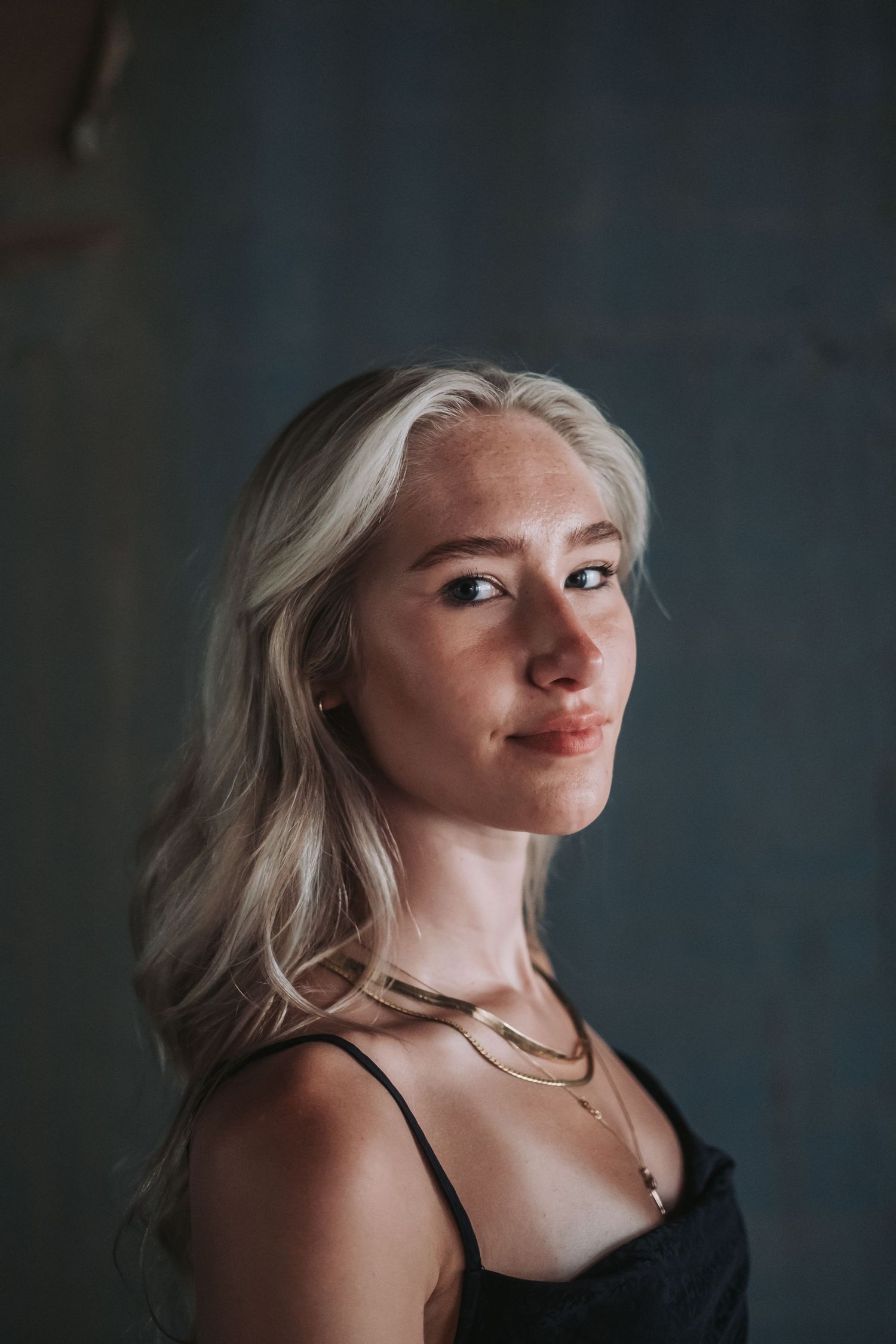 Young blonde woman wearing layered gold necklaces posing against dark textured wall.