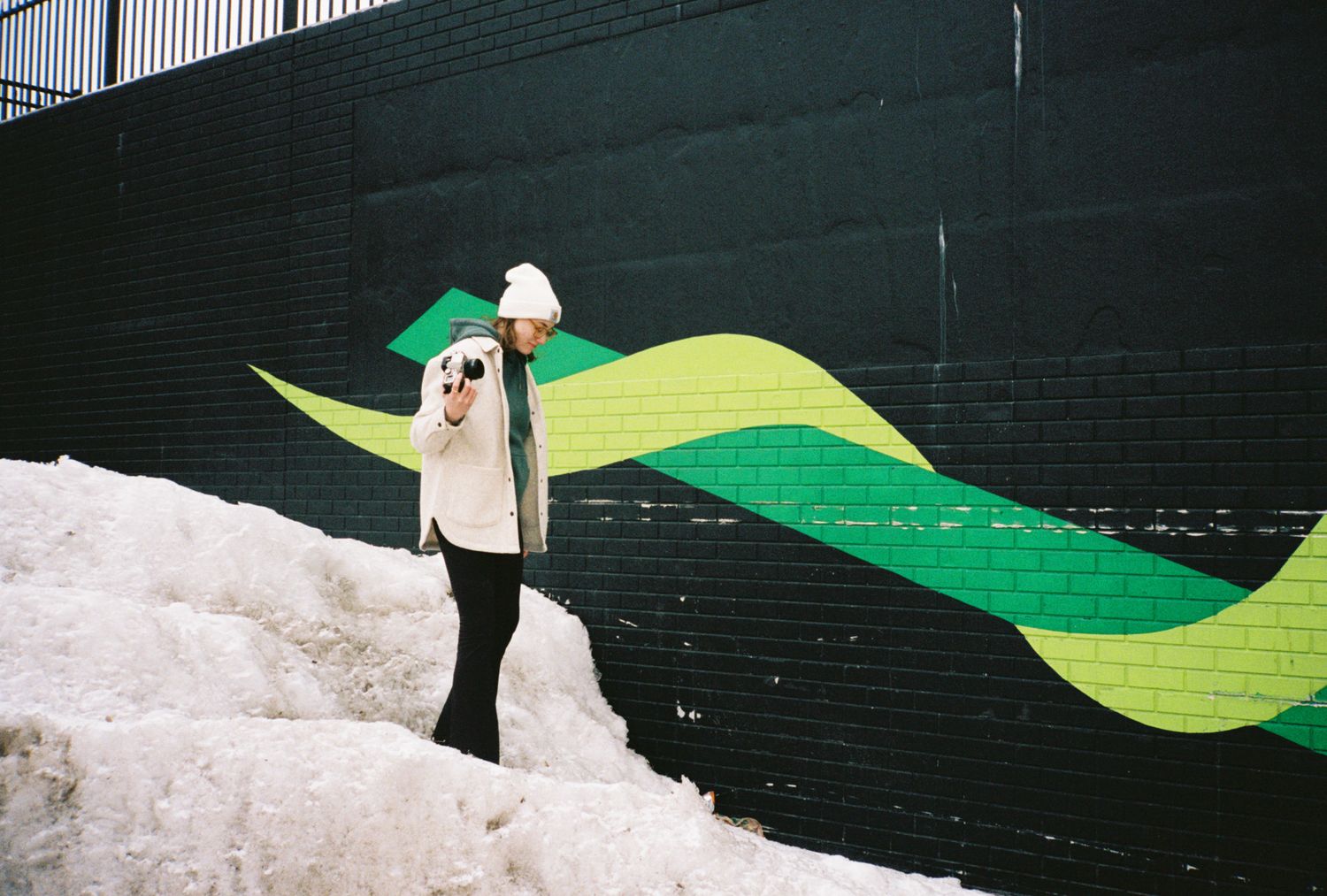 Person in white hat and jacket standing on snow beside a dark brick wall with vibrant green wave mural art.