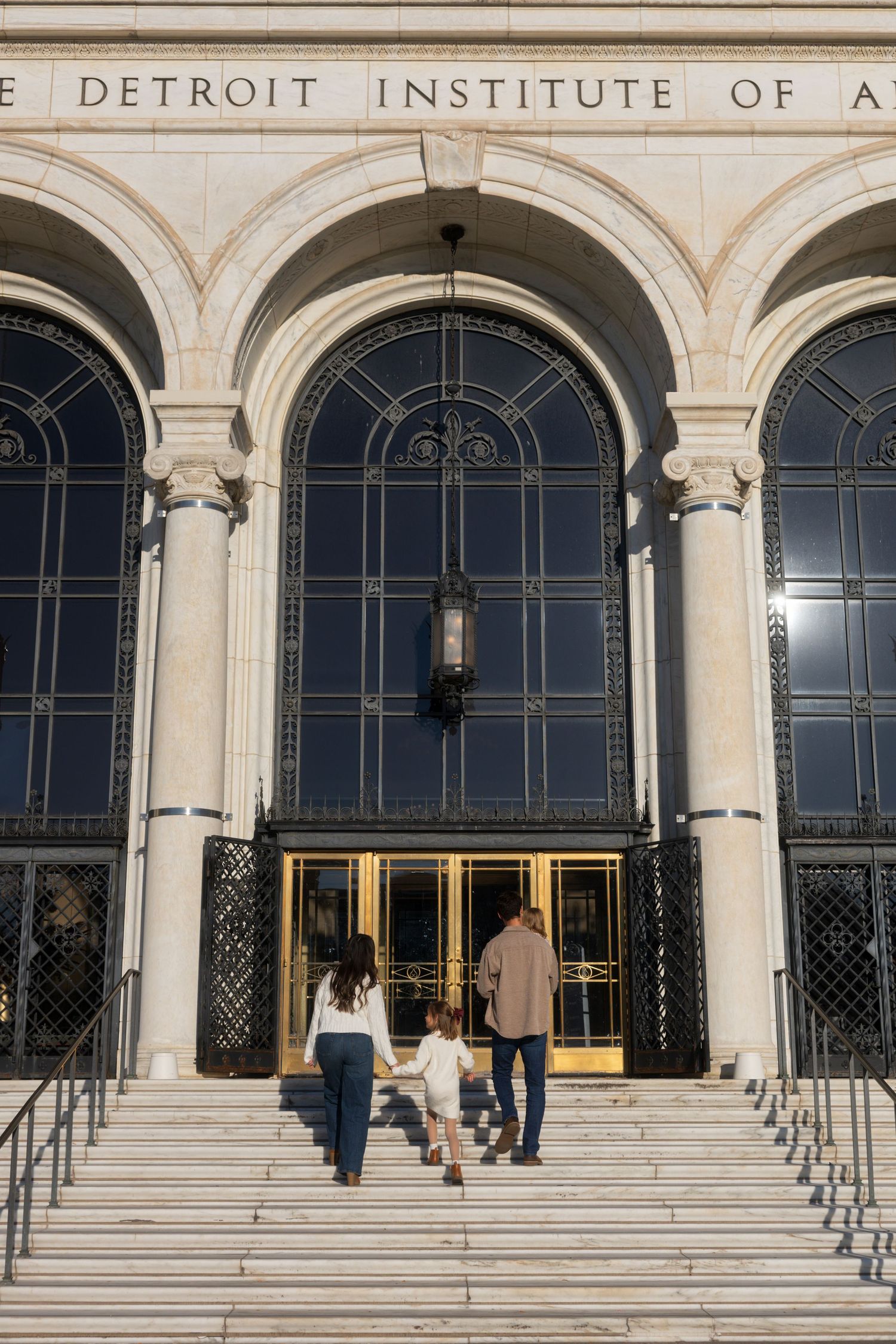 Family walking up steps to the Detroit Institute of Arts museum entrance with grand arched windows.