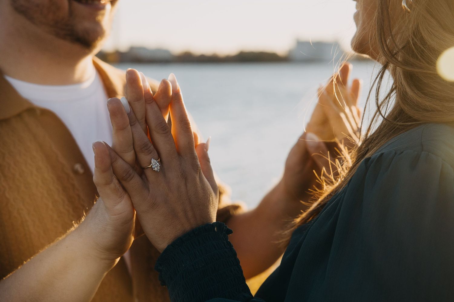 Couple pressing hands together at sunset showing engagement ring with sparkling diamond near waterfront.
