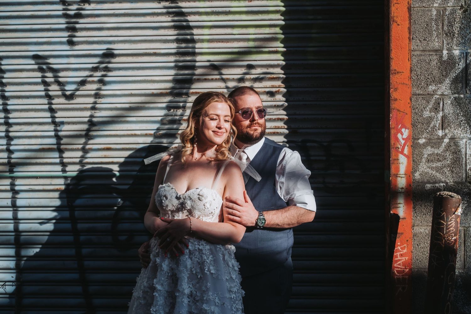 Bride in floral gown and groom in vest embrace against graffiti wall in urban wedding photo.