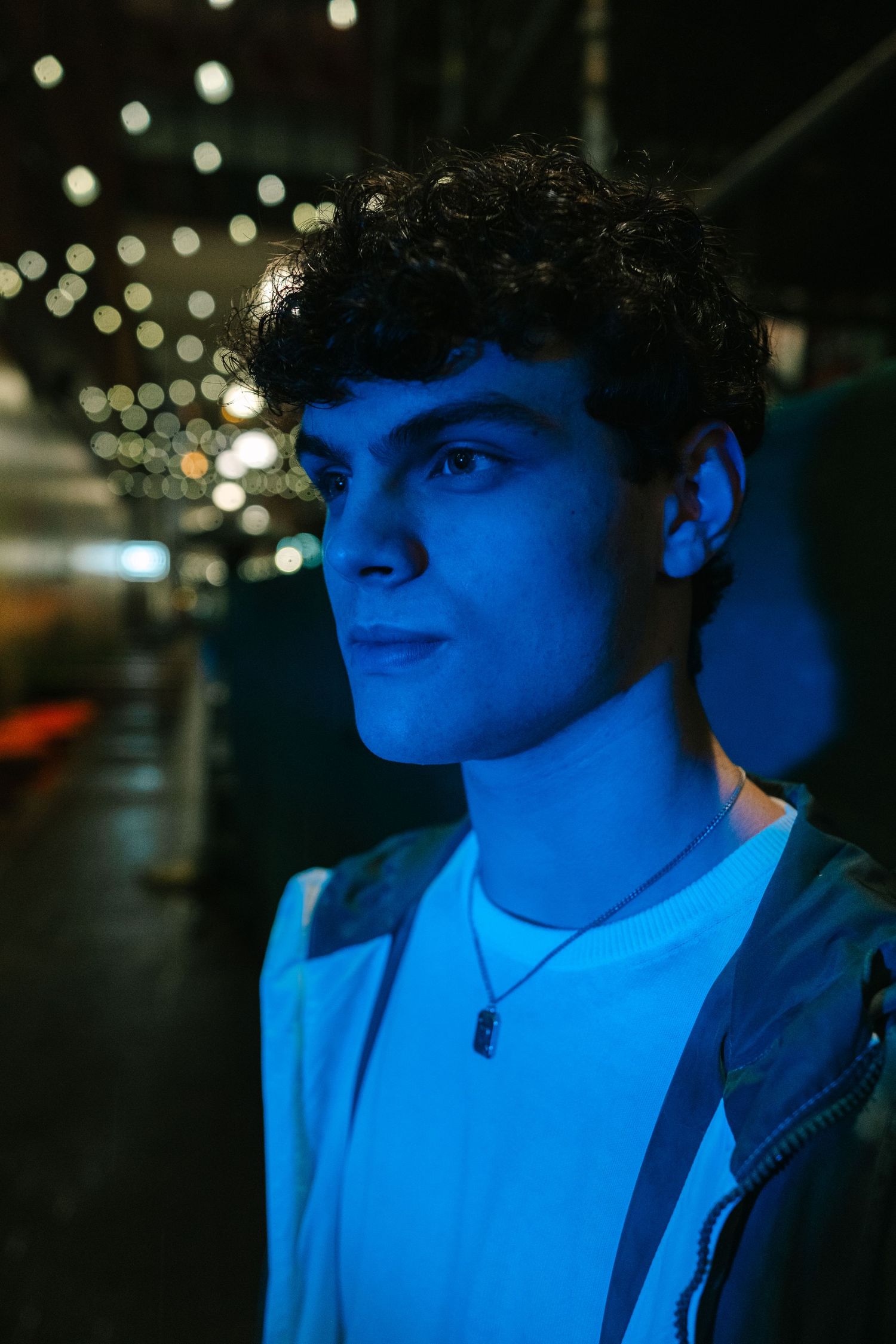 Young man with curly hair illuminated by blue neon light at night, wearing a white shirt and necklace.