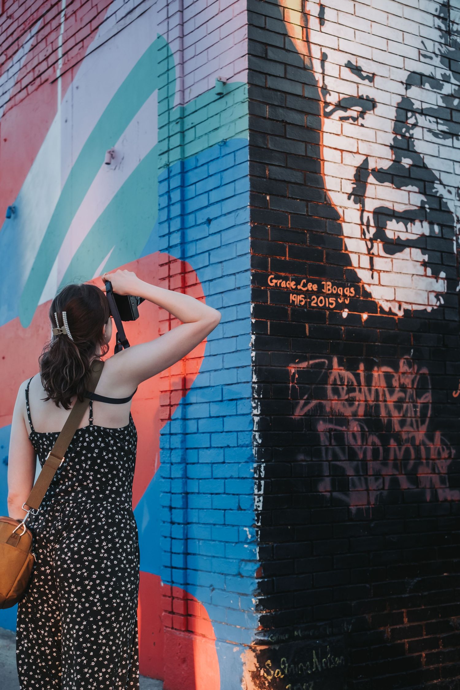Woman photographing a colorful street mural honoring Grace Lee Boggs on a brick wall.