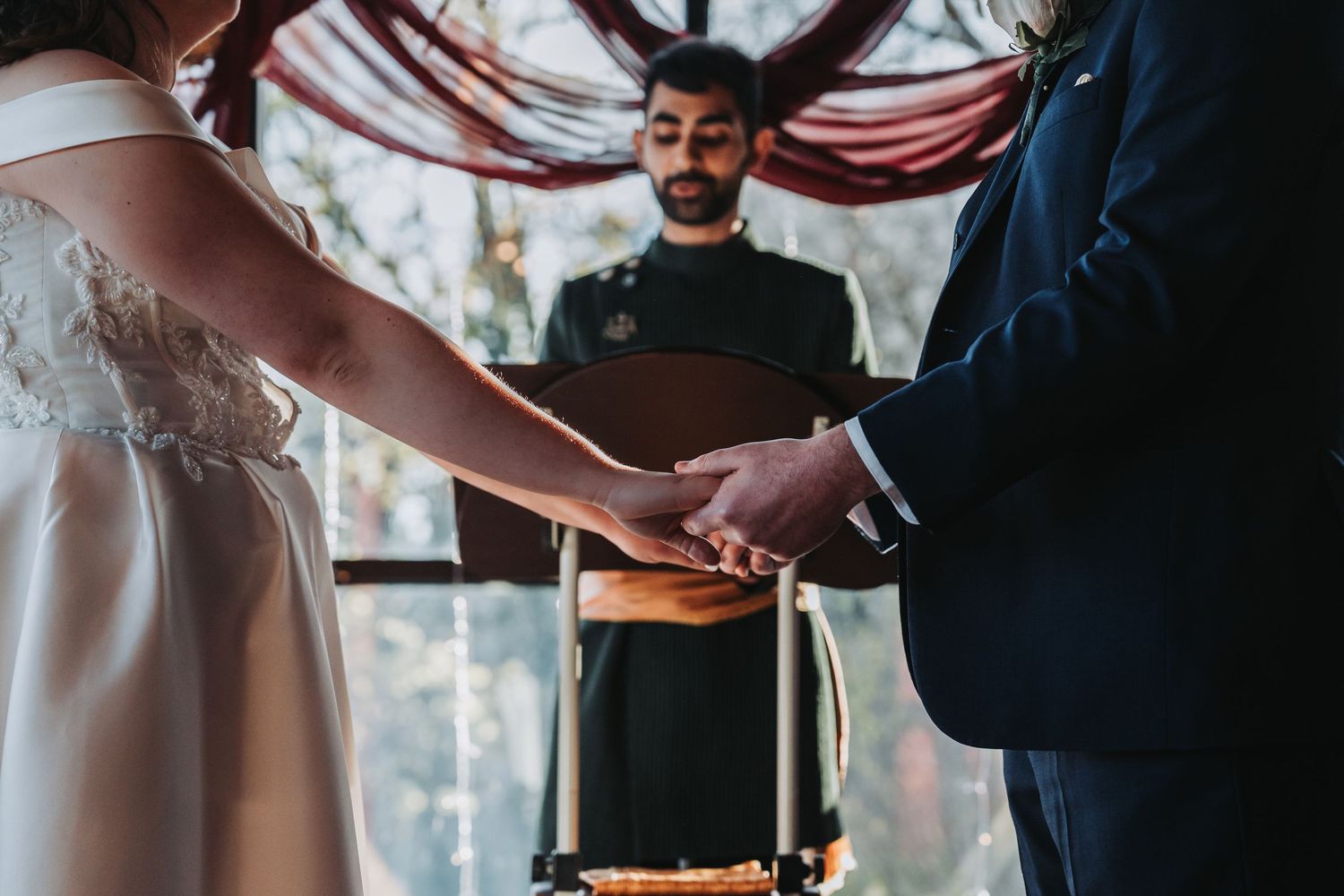 Bride and groom hold hands during wedding ceremony while officiant stands before them at decorated altar.