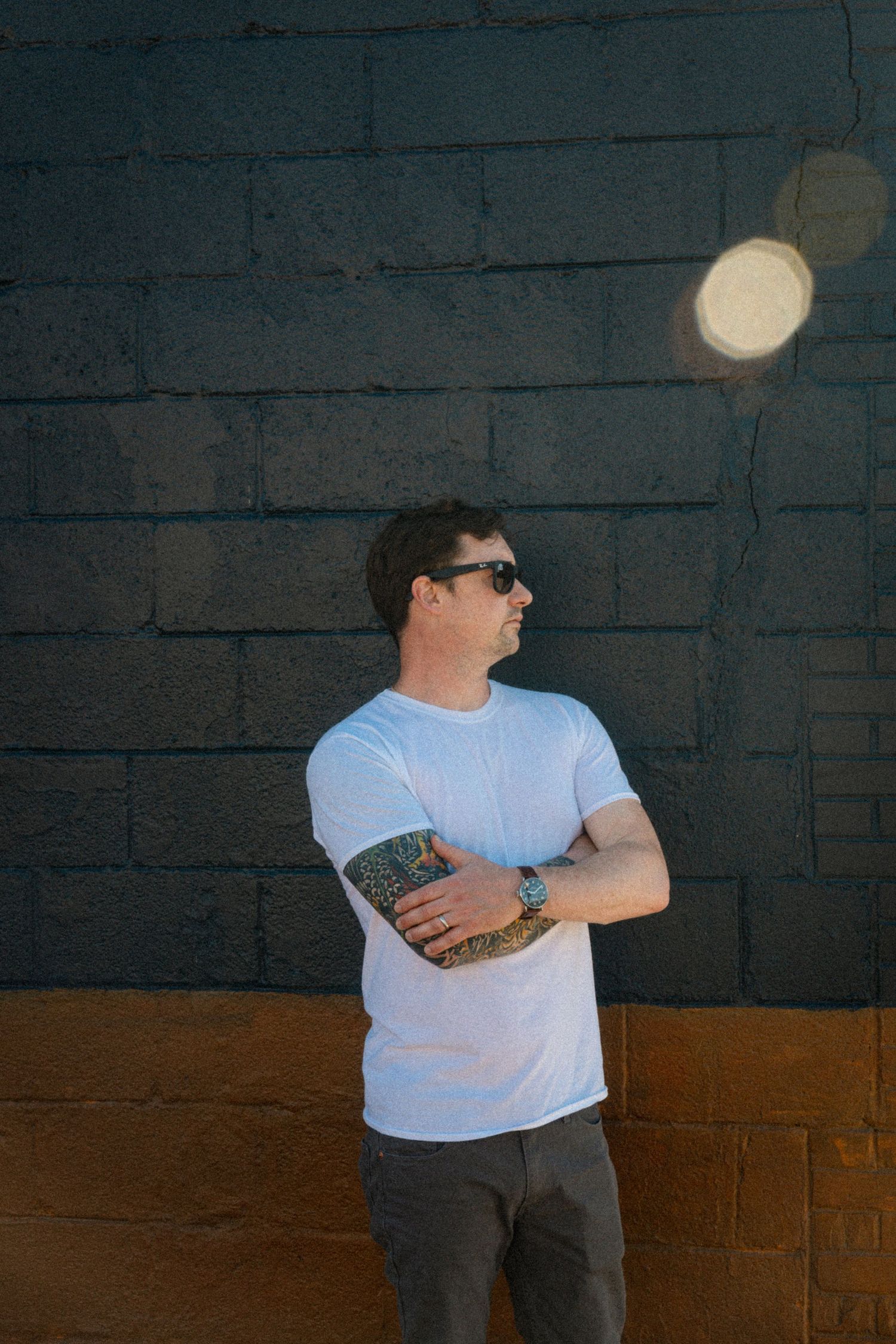 A person in a white t-shirt and dark pants stands against a dark wall with overhead lighting.