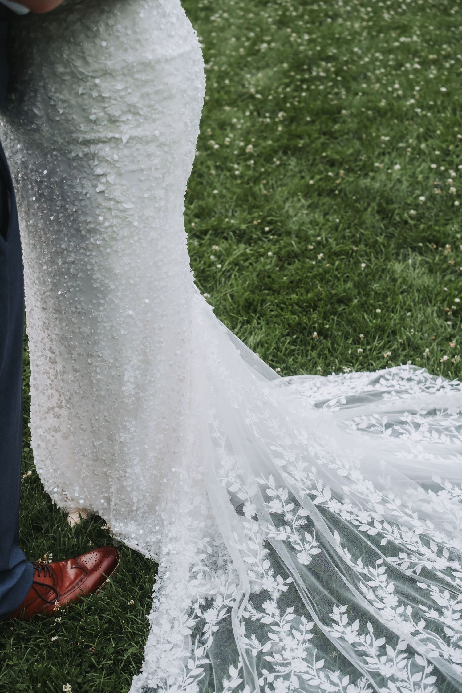 Close-up detail of an intricate lace wedding dress train trailing on green grass.