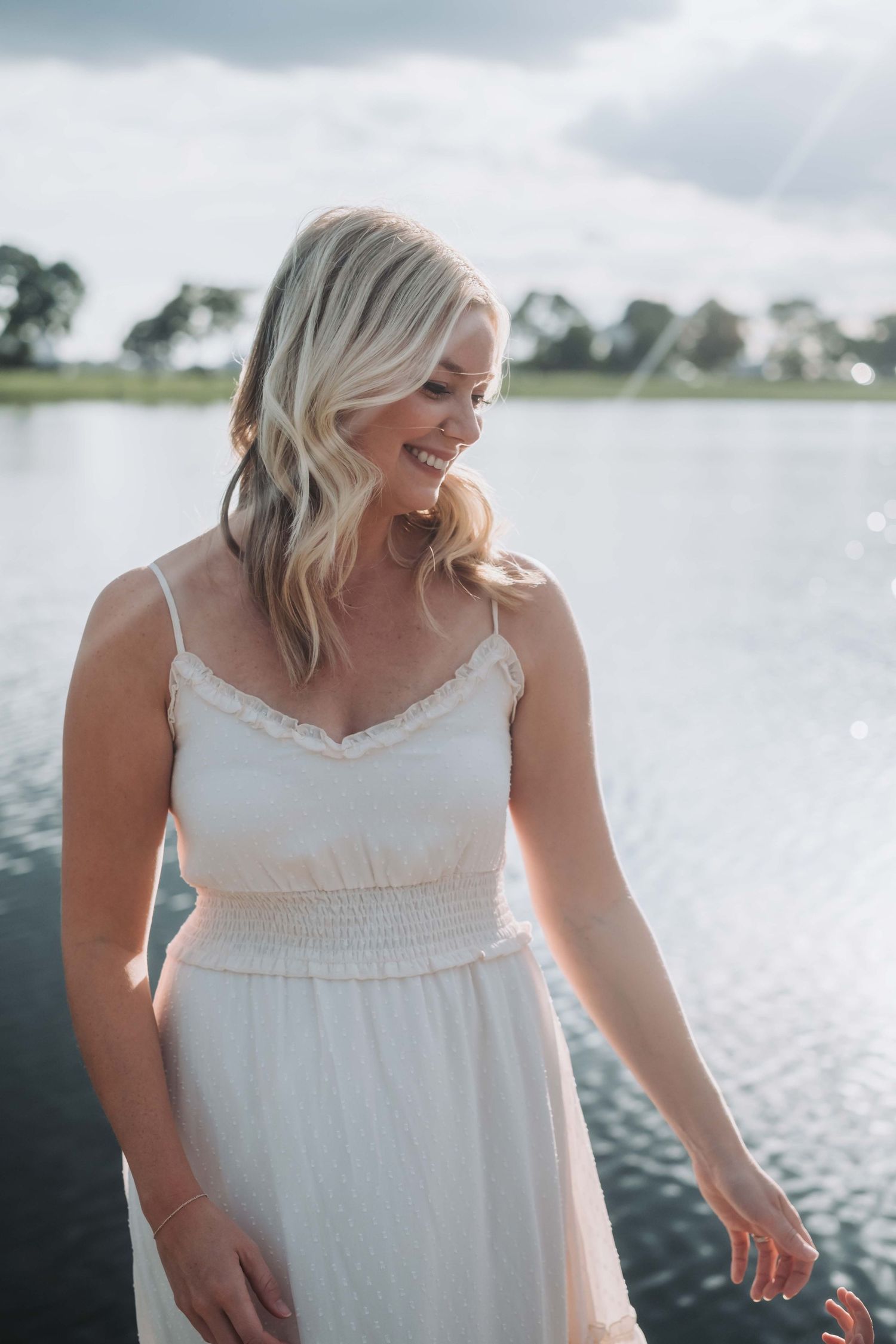 Person in white dress laughing by waterfront on cloudy day.