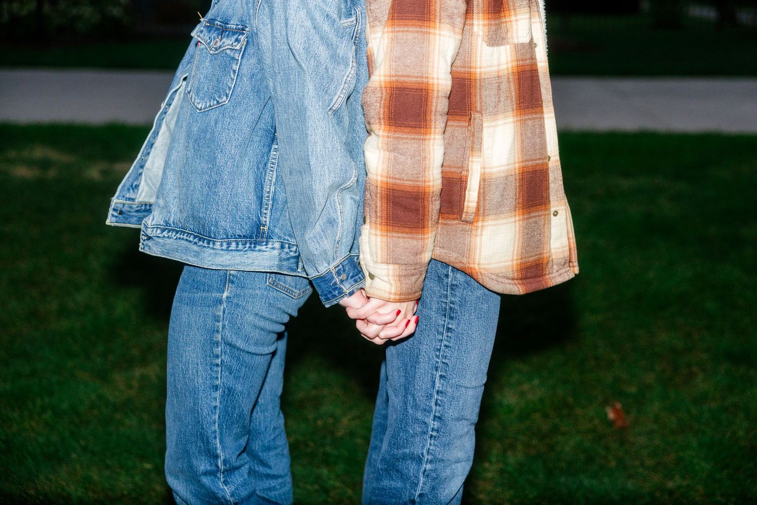 Couple holding hands wearing denim jacket and plaid shirt, standing on green grass in casual outdoor setting.