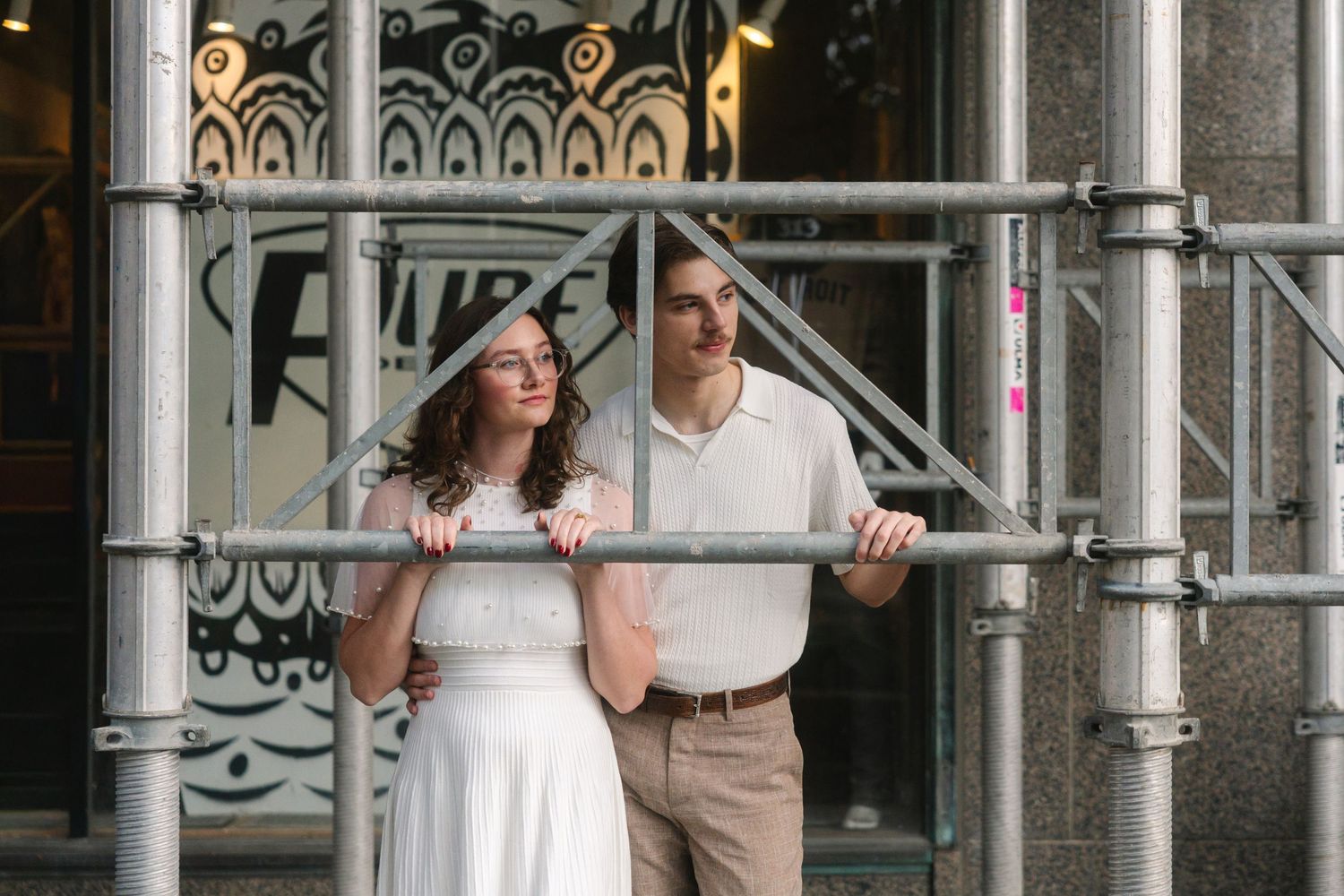 Couple in white outfits posing by metal scaffolding outside a decorated building facade.