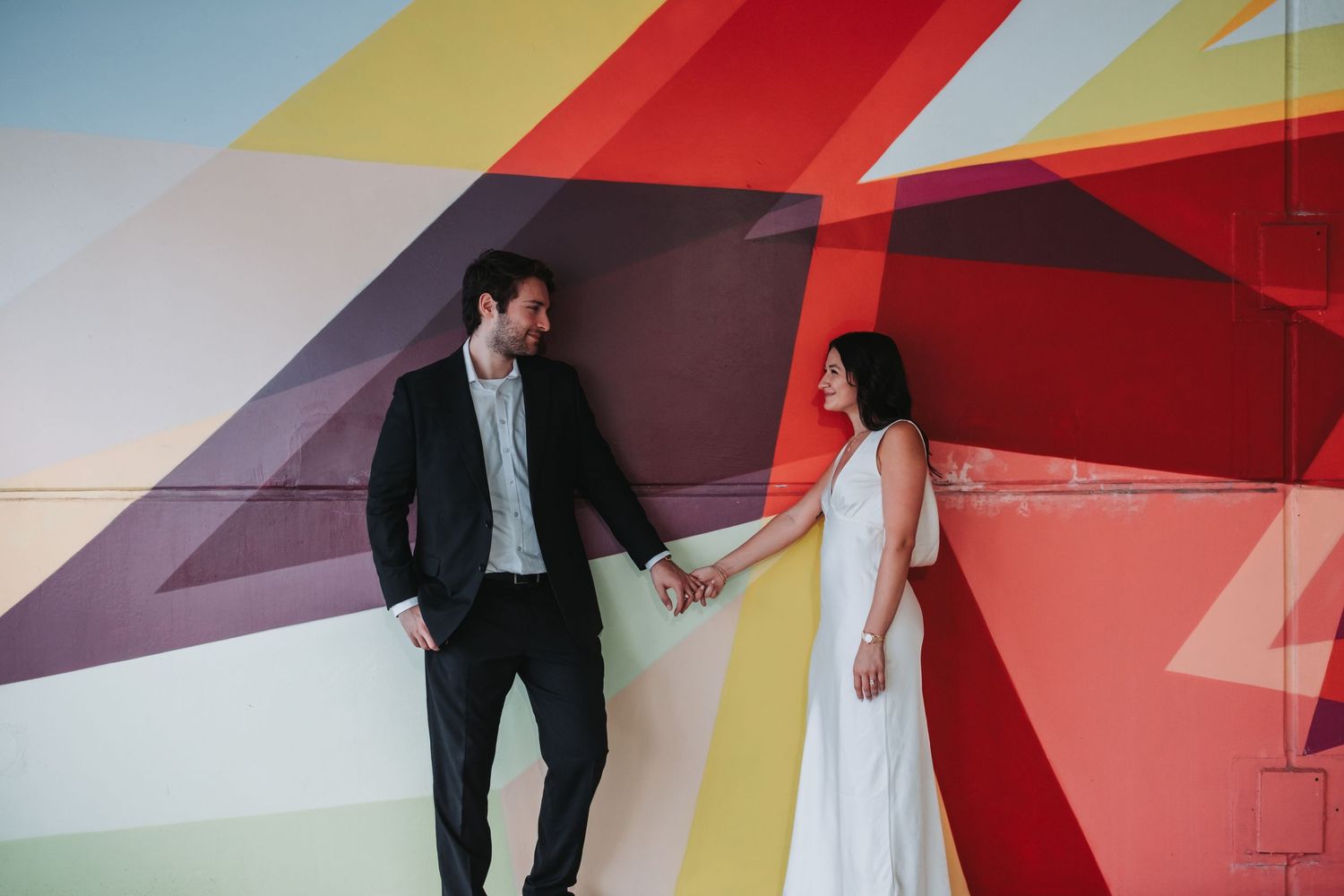 Couple holding hands in front of a colorful geometric mural, man in dark suit, woman in white dress.