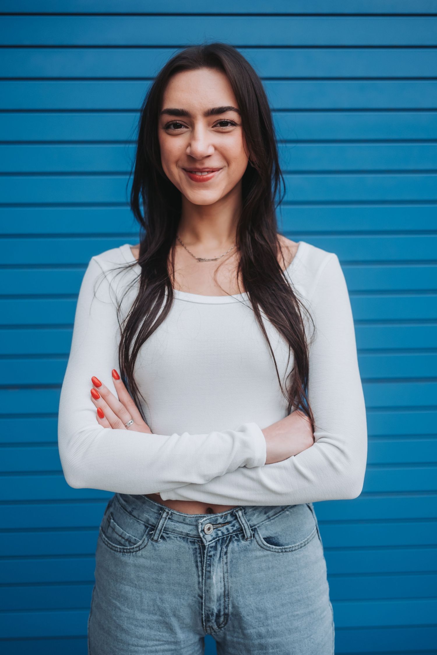 Young woman with long dark hair in white top and jeans, arms crossed, smiling against blue wall.