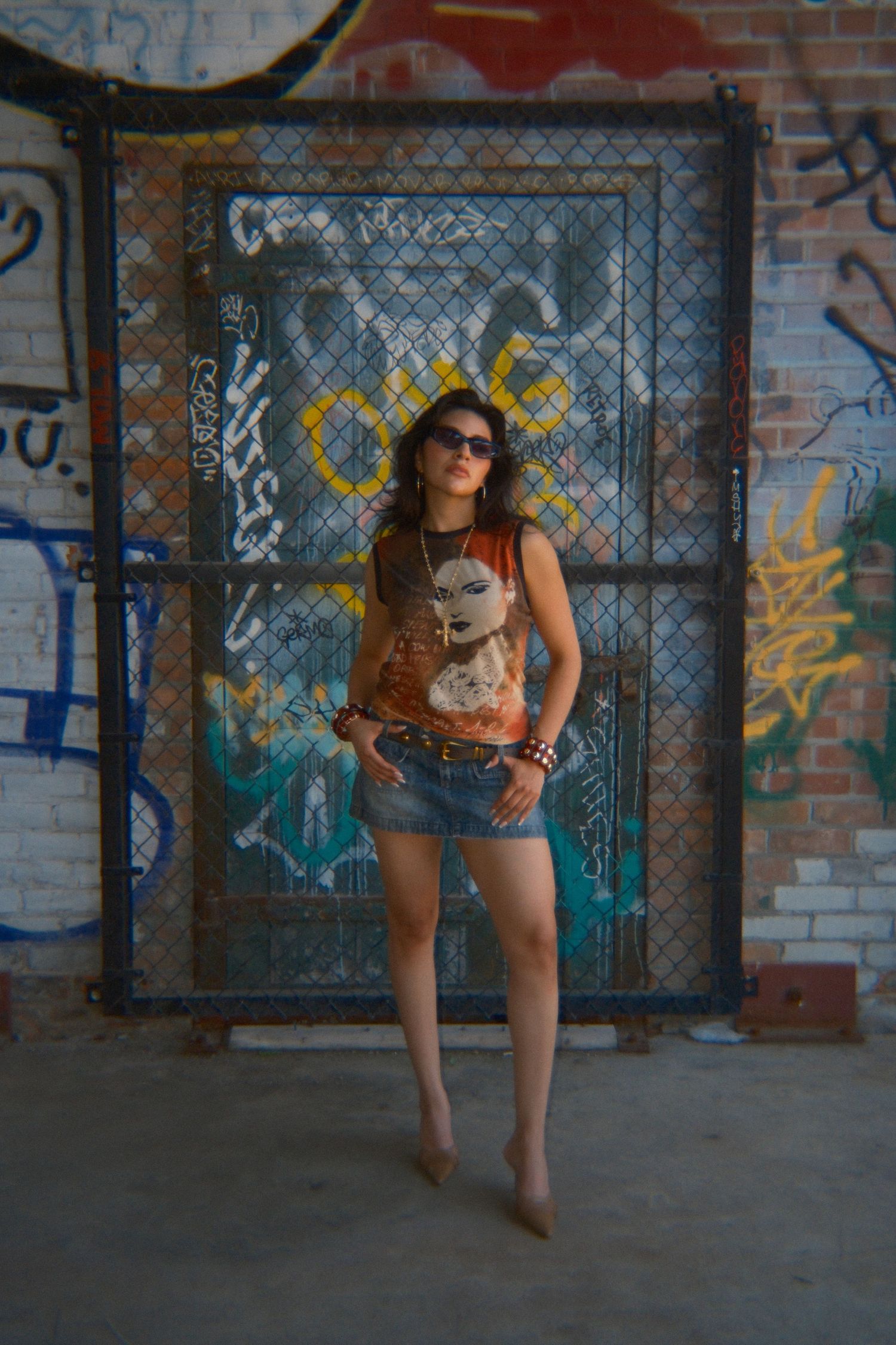 Young woman in denim shorts and graphic tee posing confidently before a graffiti-covered brick wall and chain-link gate.