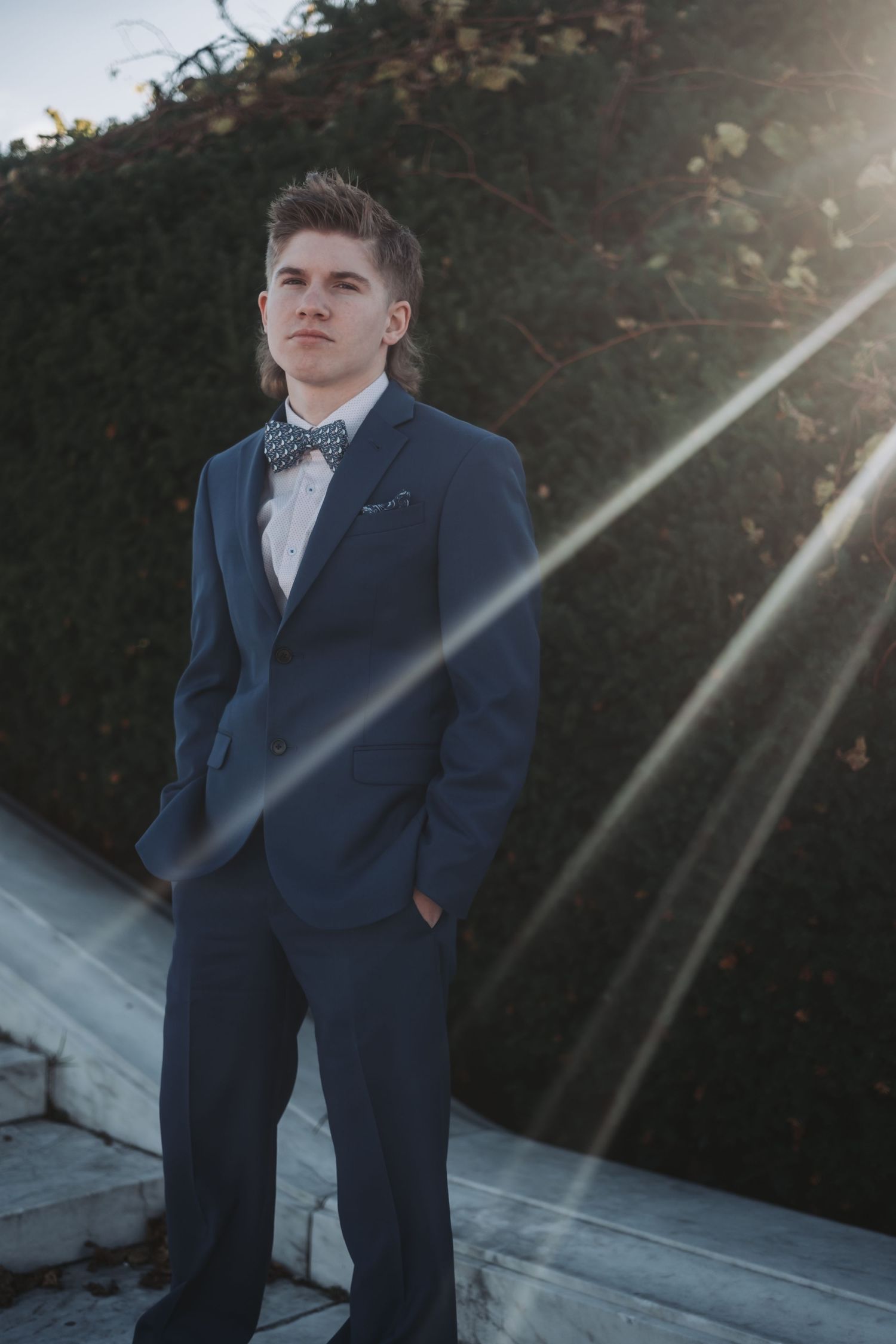 Young man in navy blue suit with bow tie stands confidently outdoors with sunlight rays and green hedge backdrop.