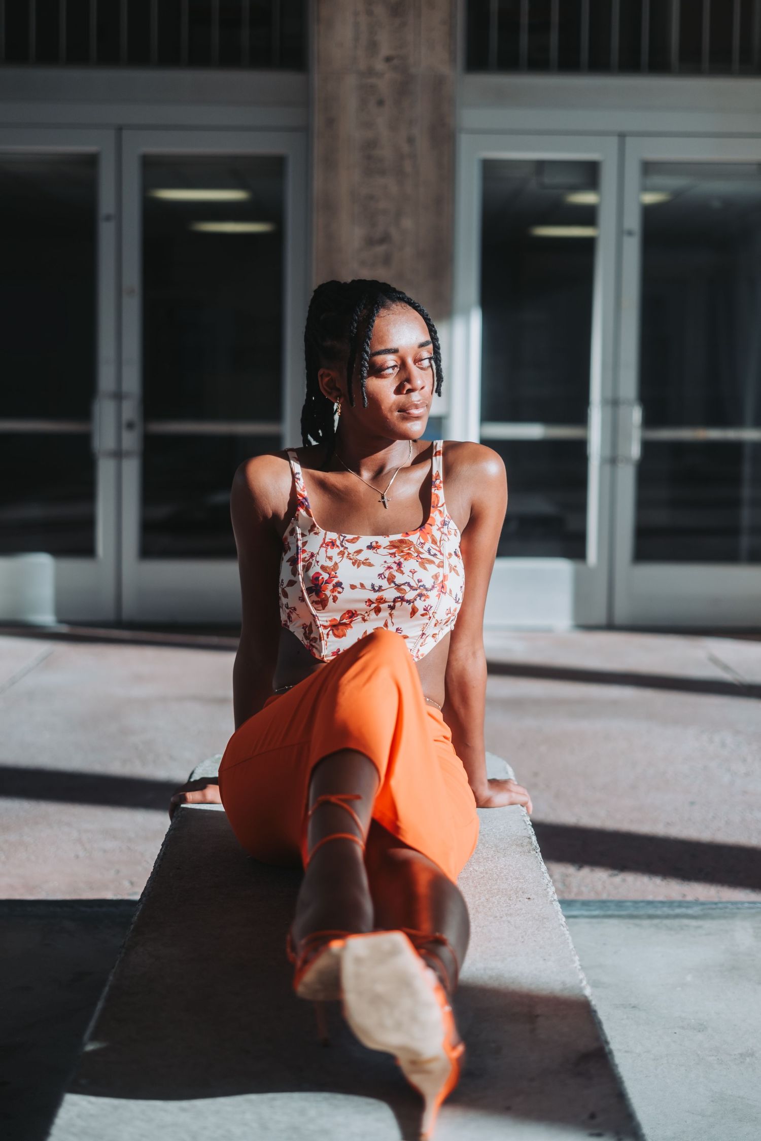 Young Black woman in floral crop top and orange pants sits on concrete ledge in sunlight outside modern building.