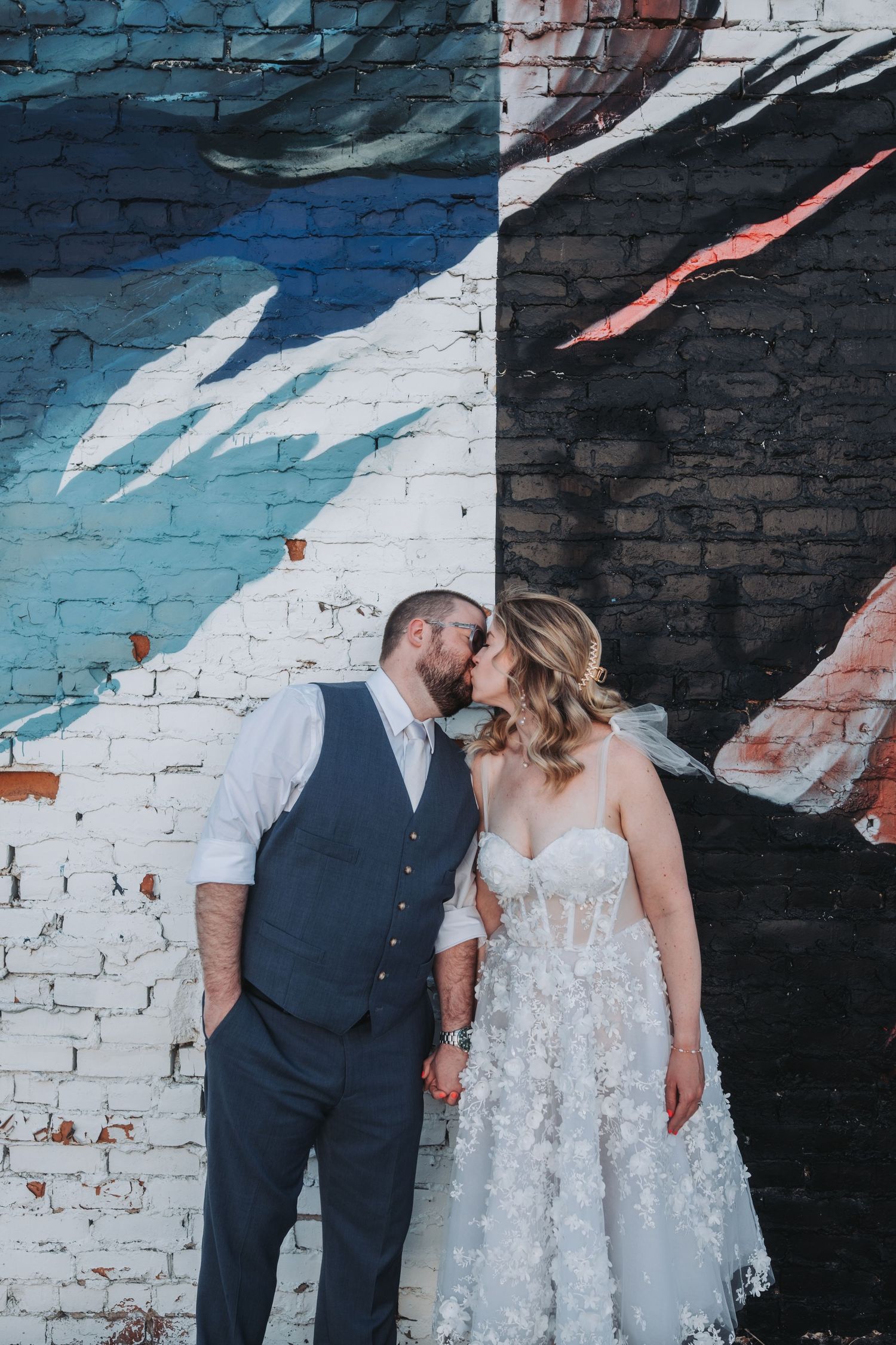 Bride and groom kissing in front of a colorful street mural on a brick wall during their wedding day.