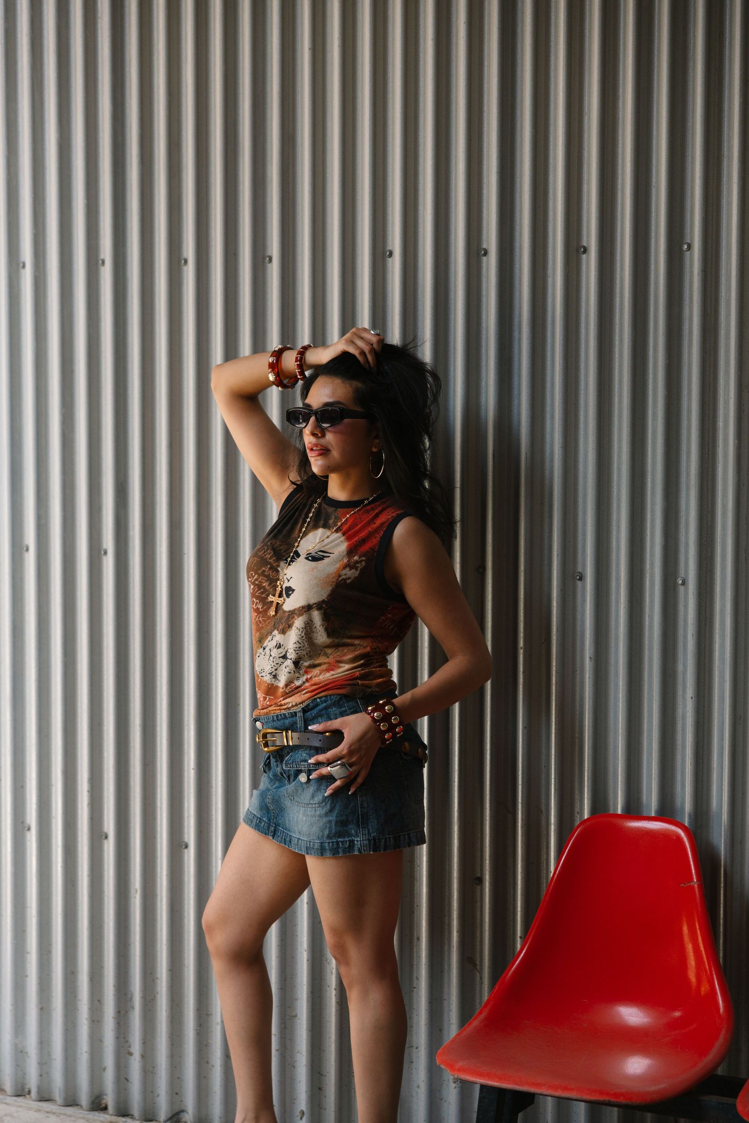 Young woman in graphic tee and denim skirt poses against corrugated metal wall beside a red chair.