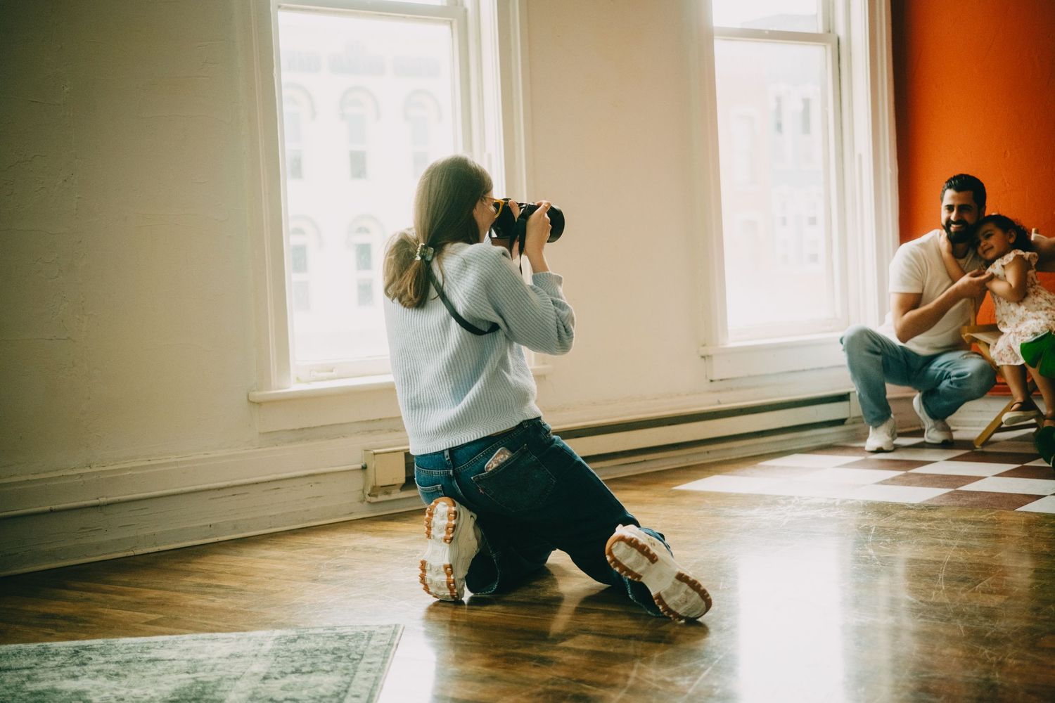 Photographer kneeling on hardwood floor capturing photos of smiling man and child near bright windows indoors.