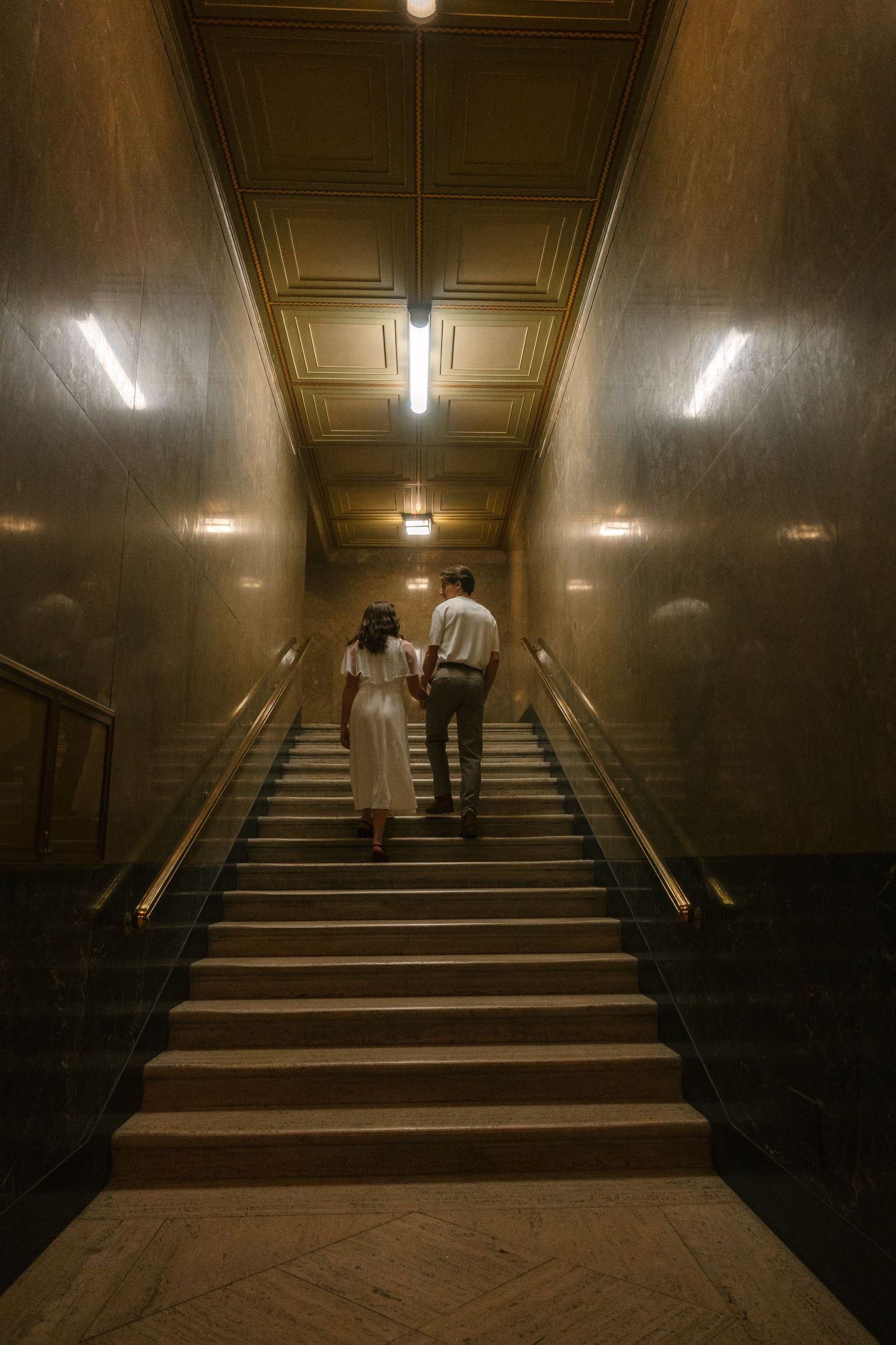 Two figures ascending dimly lit stairs in a vintage building with dramatic shadows and warm lighting.