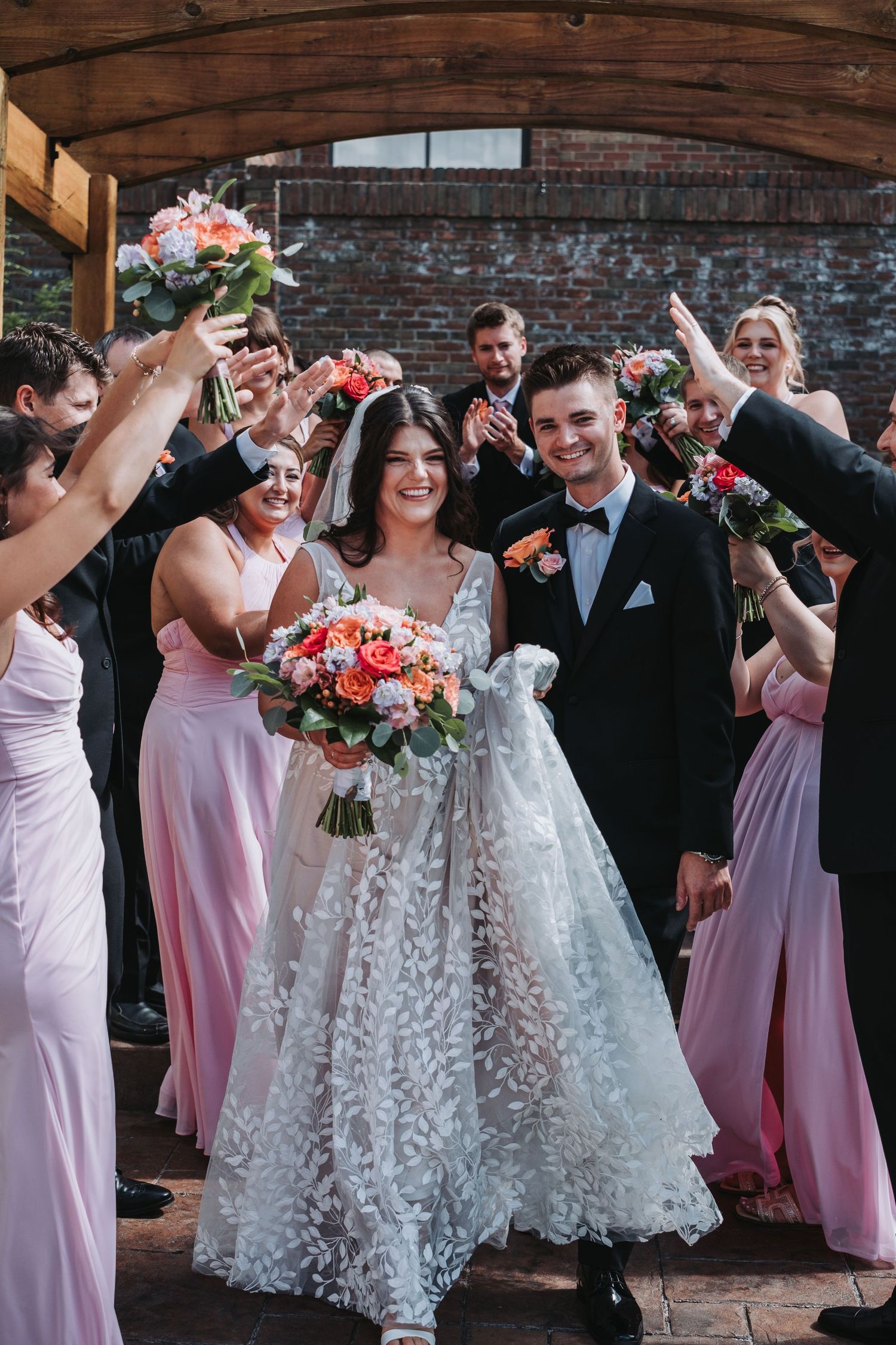 Wedding party in pink bridesmaid dresses and black tuxedos celebrate together against a brick wall backdrop.