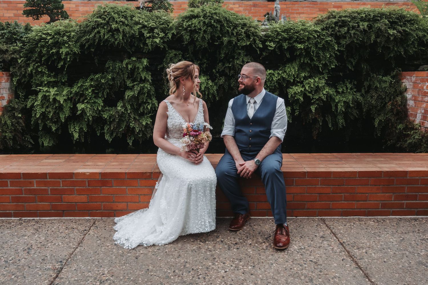 Couple sitting on brick wall bench surrounded by green foliage during wedding celebration.