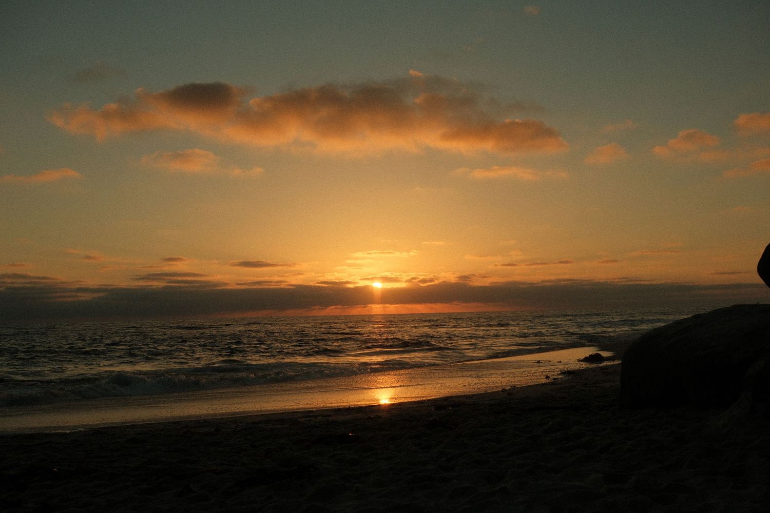 Golden sunset over calm ocean waves with dramatic clouds and sandy beach shoreline at dusk.