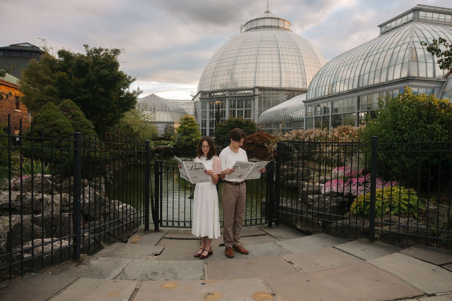 Couple exchanging vows in front of a grand glass greenhouse botanical garden with lush greenery and iron gates.