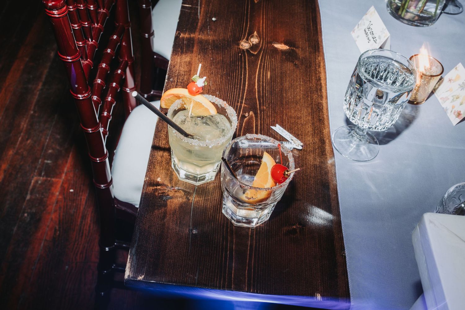 Two cocktails with citrus garnishes on a wooden bar table, surrounded by glassware in a dimly lit venue.