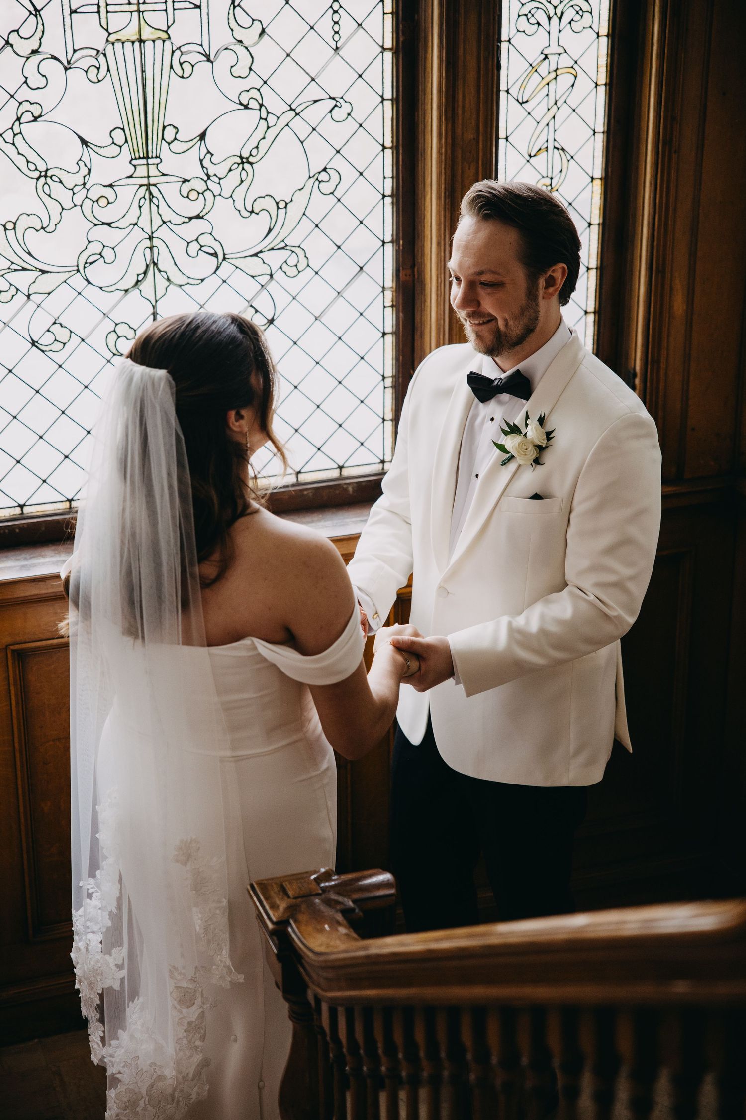 Groom in white tuxedo smiling at bride during wedding ring exchange near ornate stained glass window.