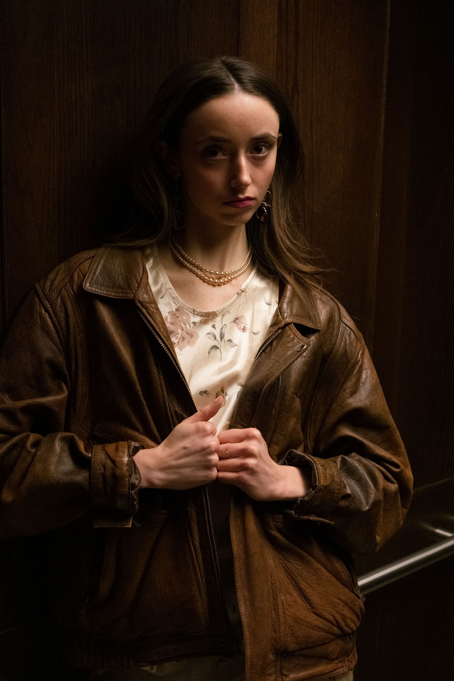 Young woman in brown leather jacket and gold necklace leaning against wooden wall with serious expression.