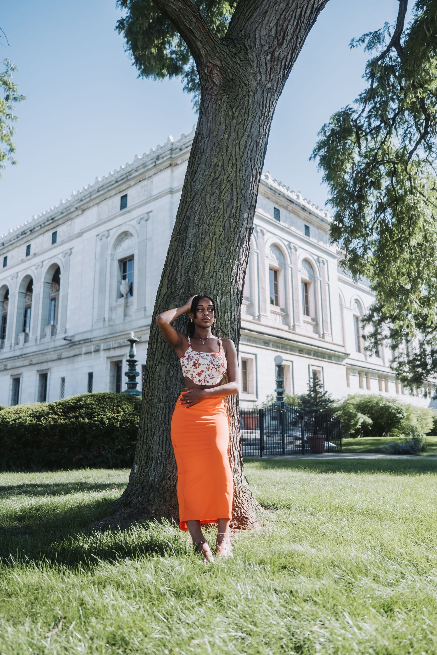 A woman in an orange skirt and floral top leans against a large tree in a park near a classic white building.