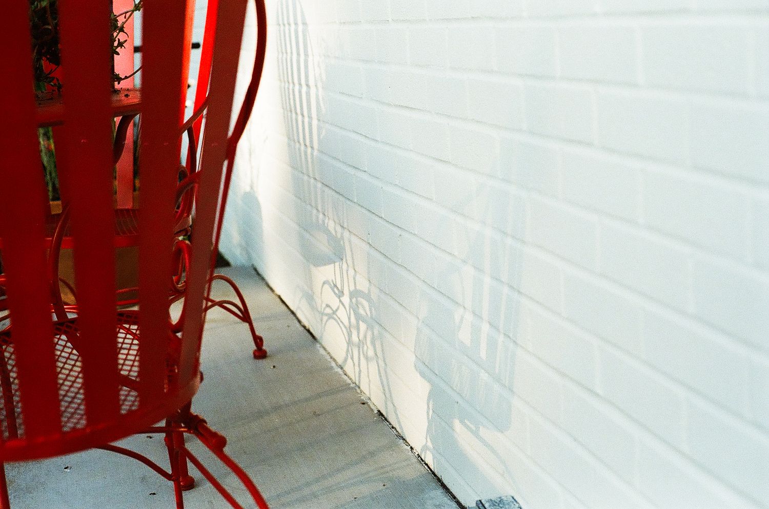 Red metal patio chair casting shadow on white brick wall outdoors on concrete ground.