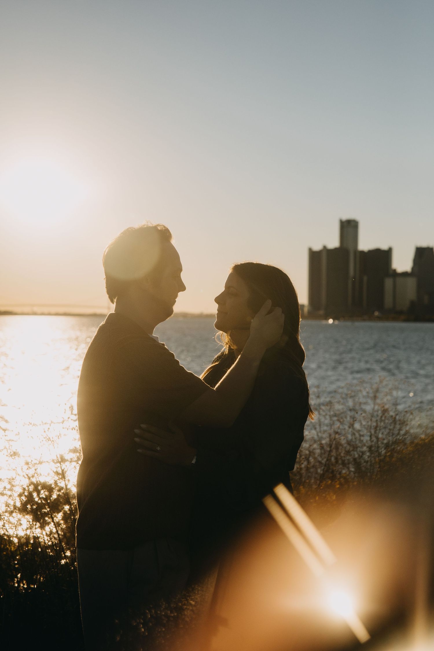 Two silhouetted figures share an intimate moment against a glowing sunset by the waterfront with city buildings in background.