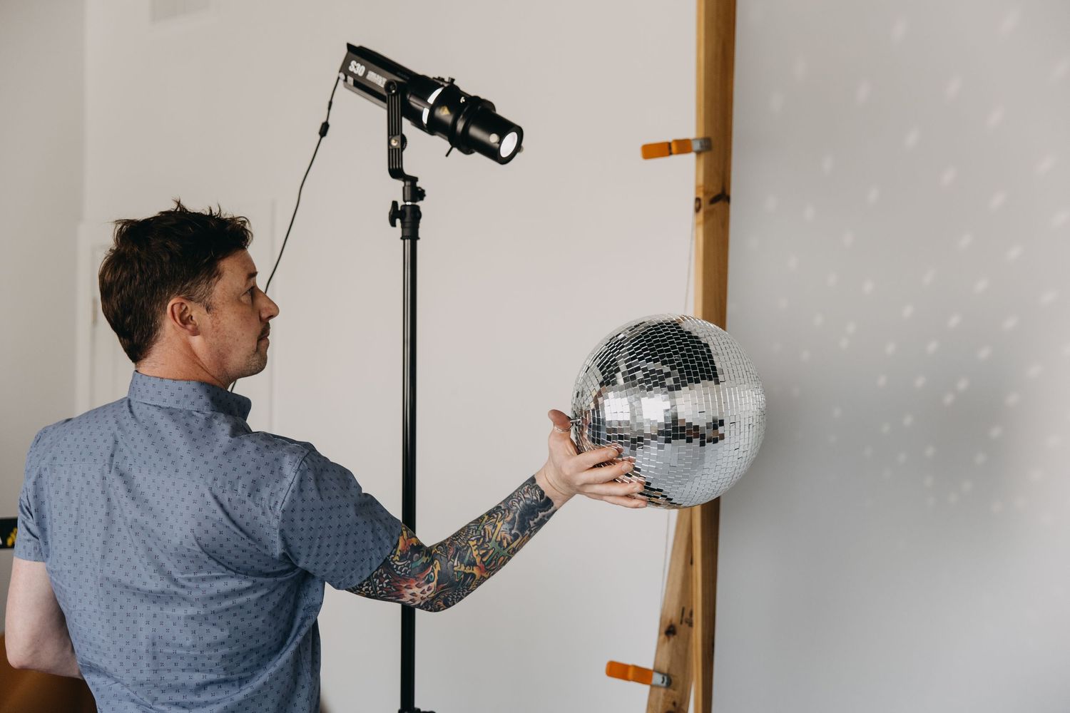 Tattooed man holding a disco ball near a photography studio light on a tripod against a white wall.