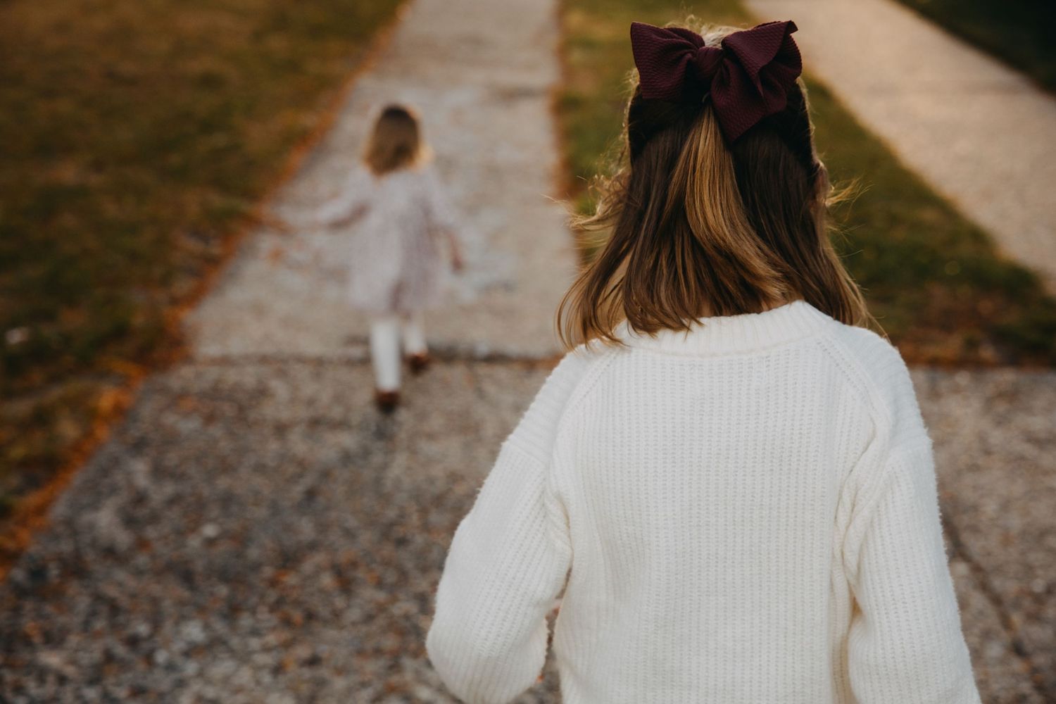 Two young girls in white sweaters walk down a path, the closer one wearing a burgundy bow in her hair.