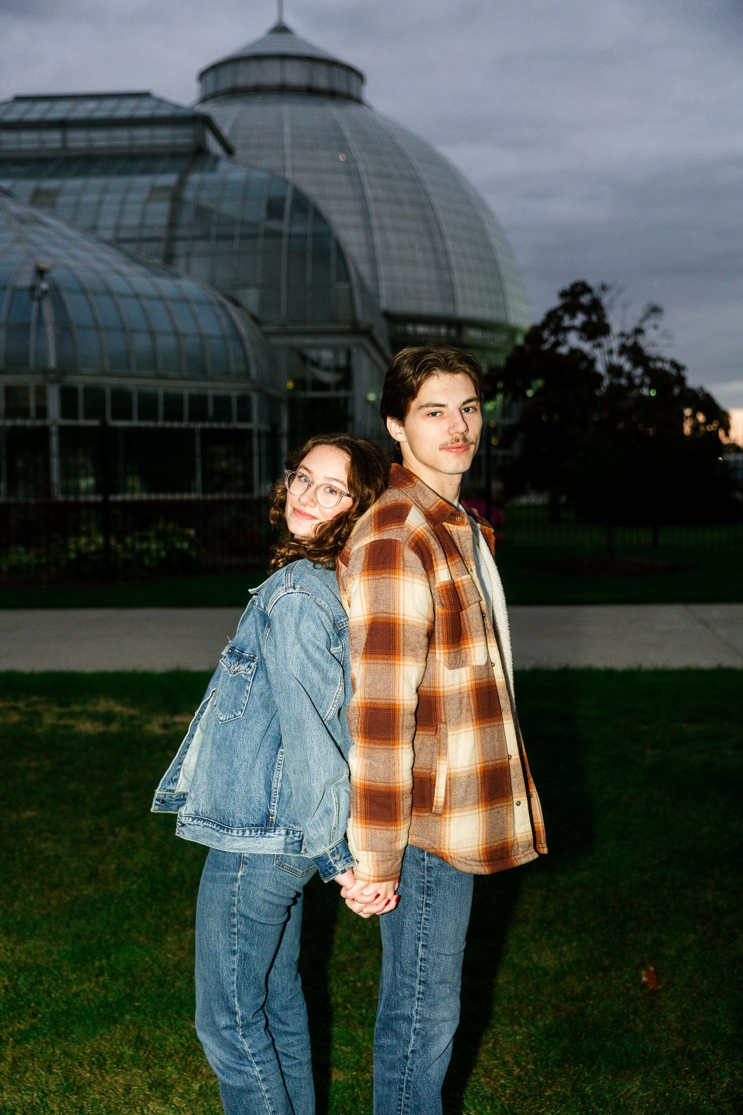 A young couple holds hands and poses in front of a large glass greenhouse dome in a park at dusk.