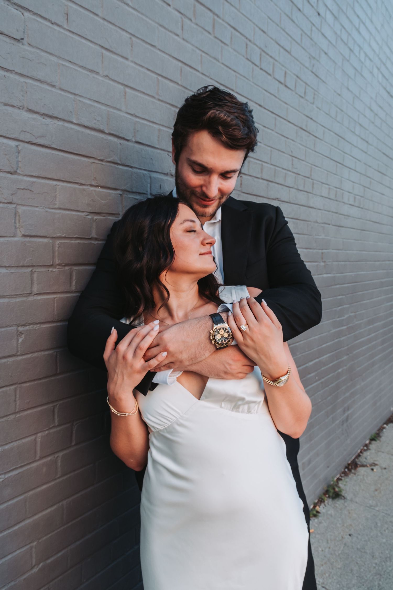 Couple embracing against gray brick wall, bride in white dress, groom in black suit, sharing a romantic moment.
