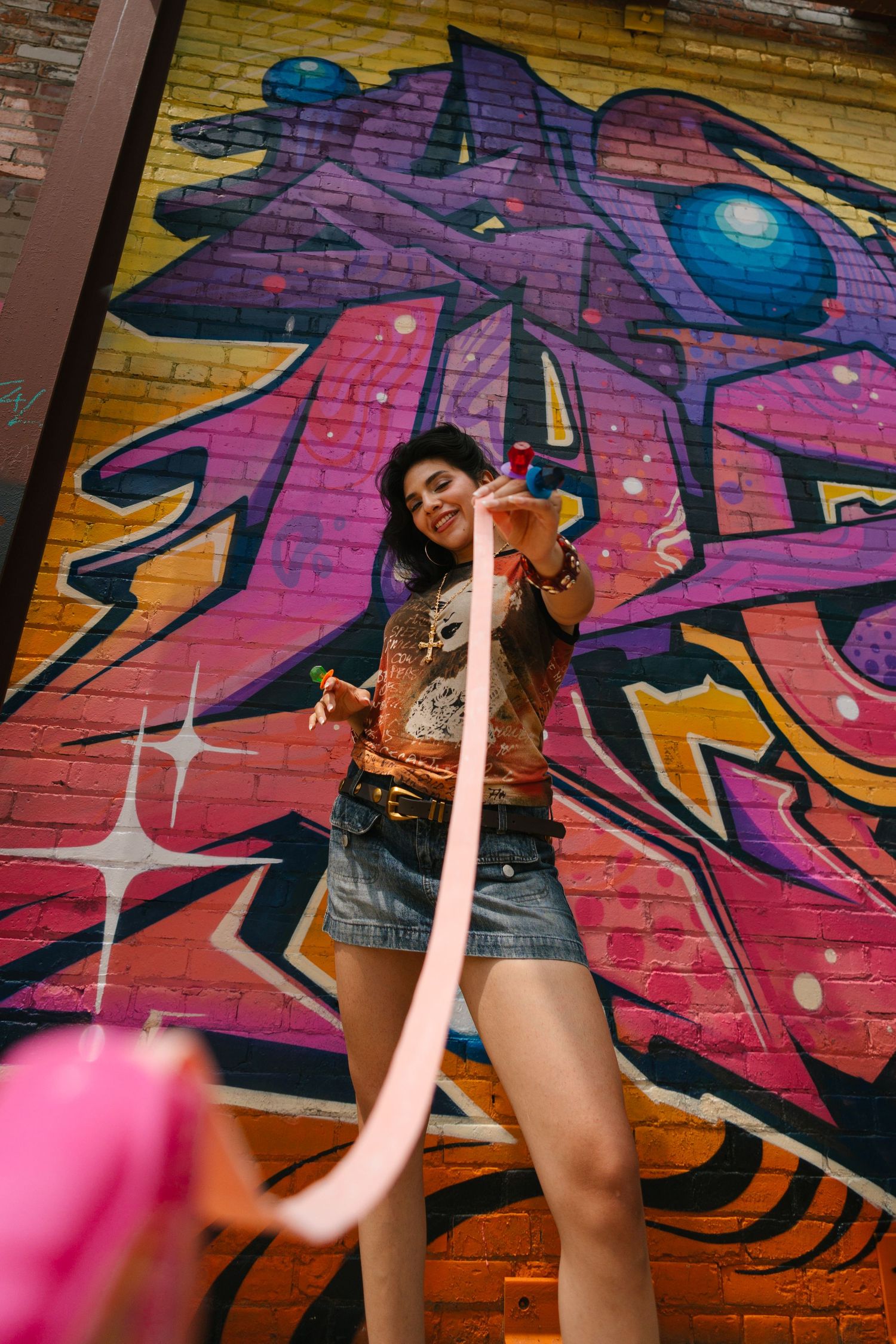 Woman in denim shorts poses with pink ribbon in front of colorful graffiti wall.