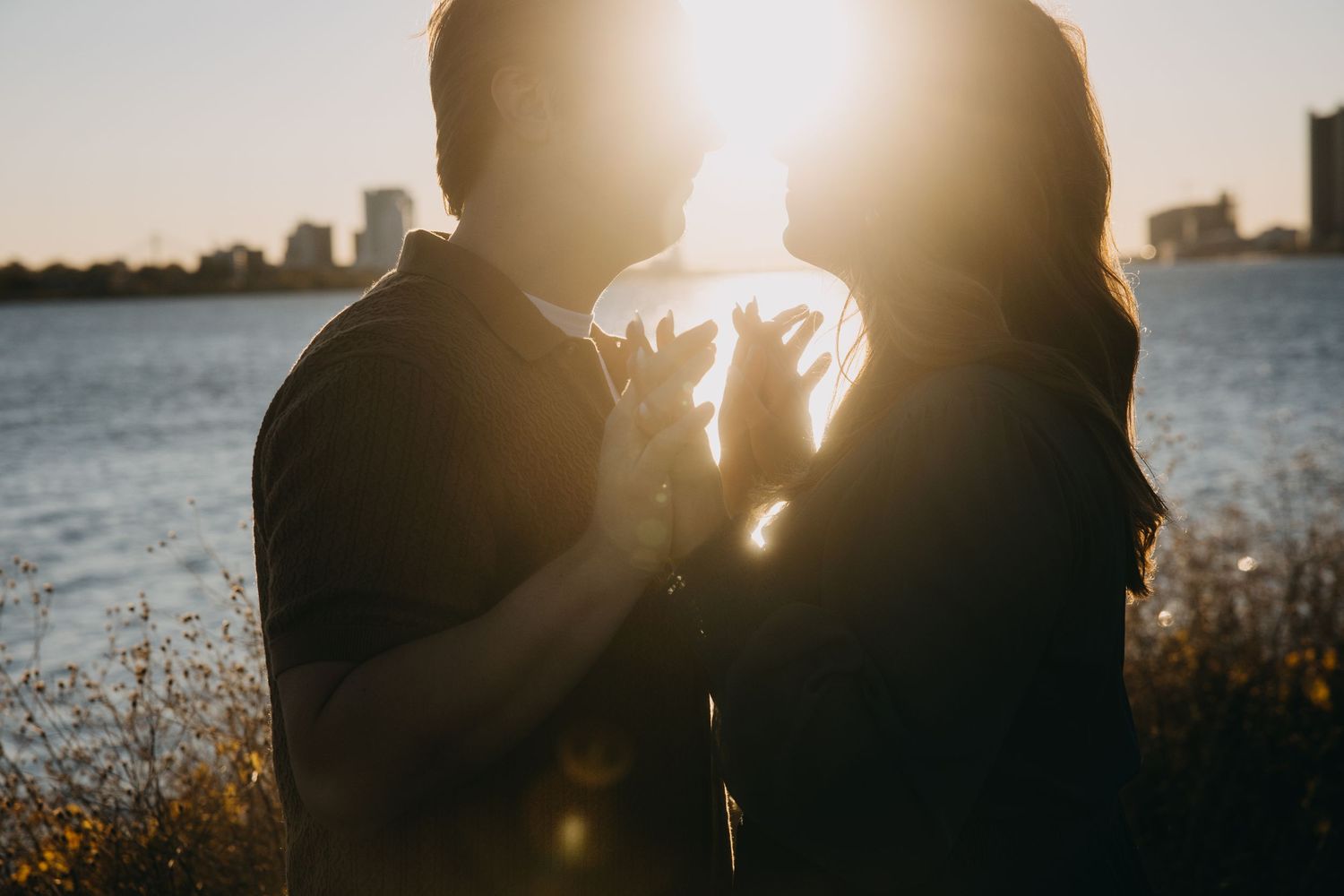 Couple sharing a romantic moment at sunset by the water with golden backlight and city skyline in the background.