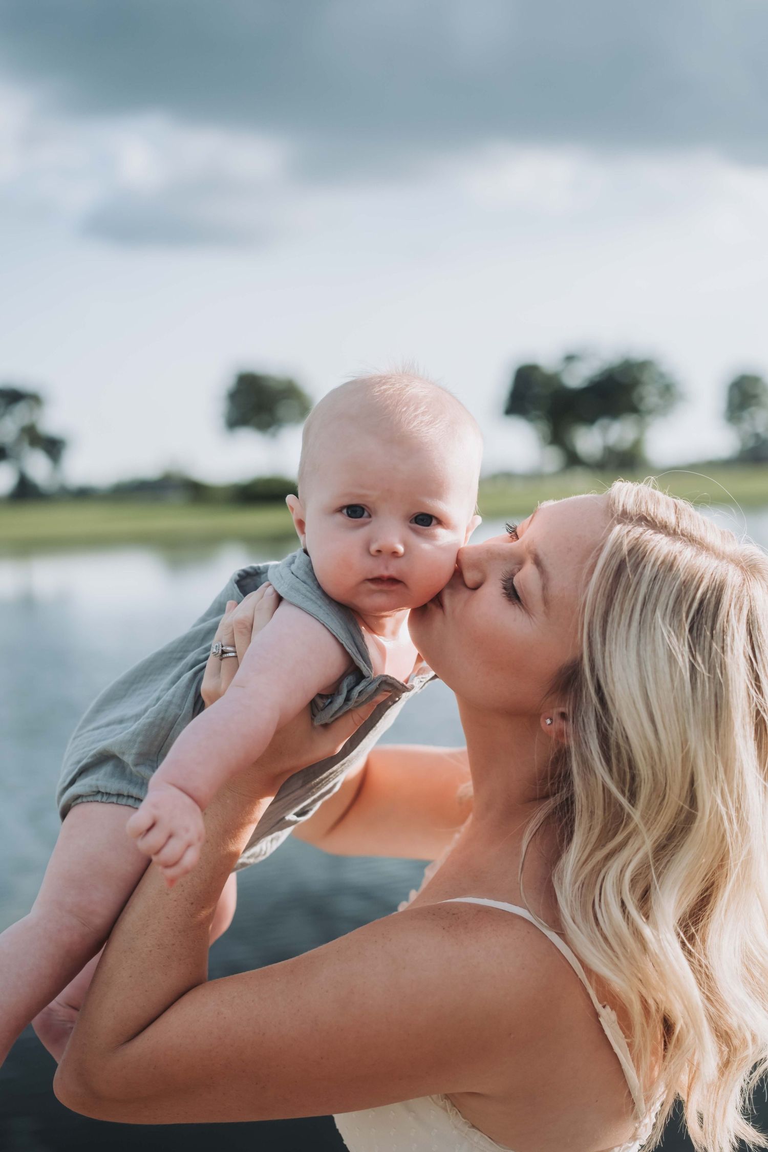 Mother and baby sharing a tender moment against a cloudy sky by the water.