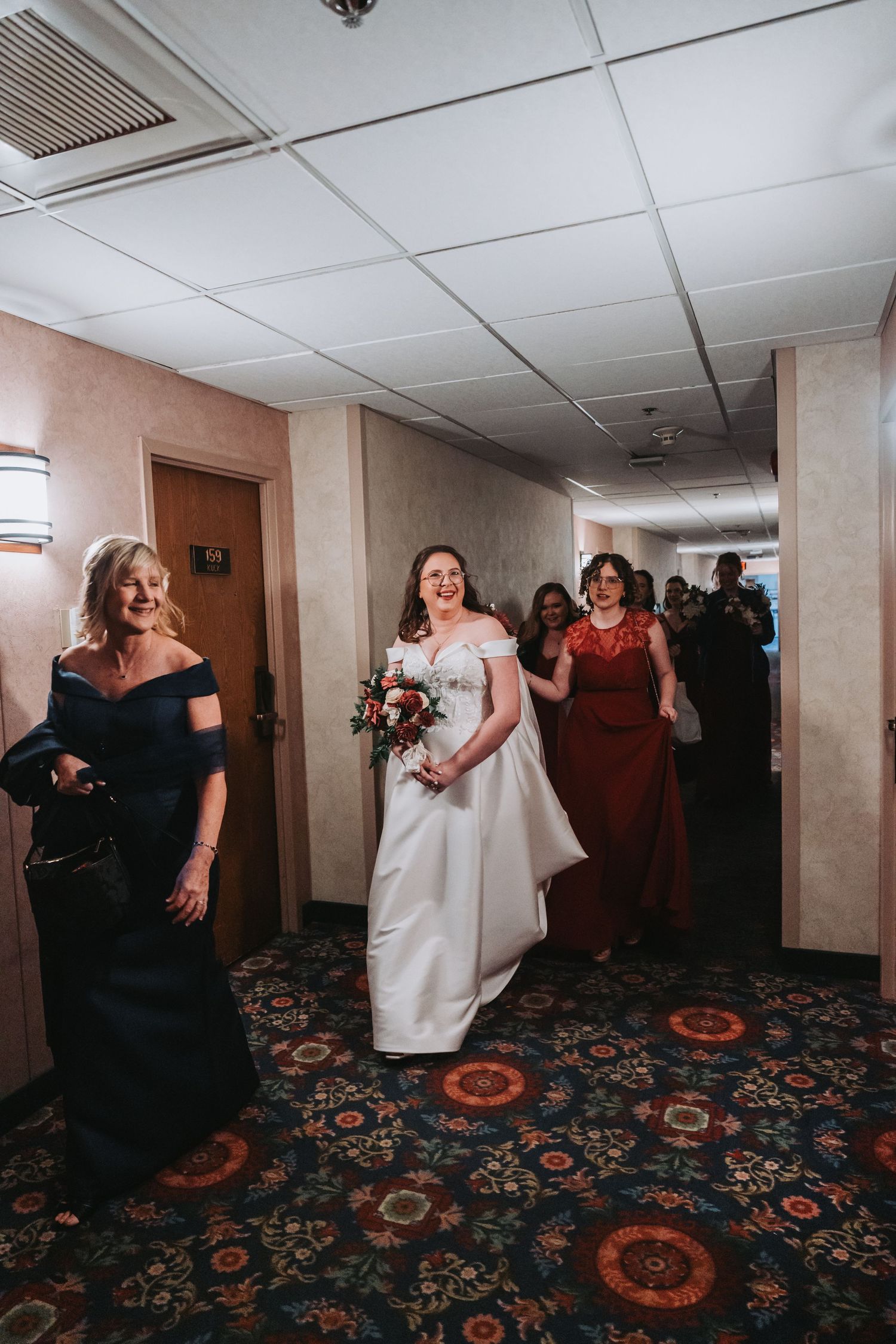 Wedding party walking down a hotel hallway with bridesmaids in dark gowns and a bride in white.