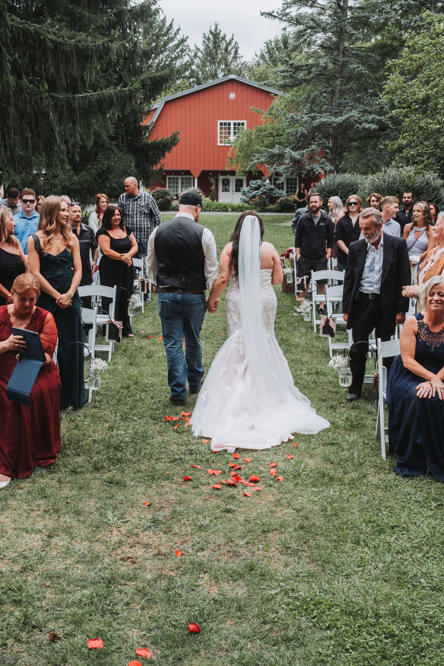 Outdoor wedding ceremony with rose petals scattered along the aisle and guests seated on white chairs.